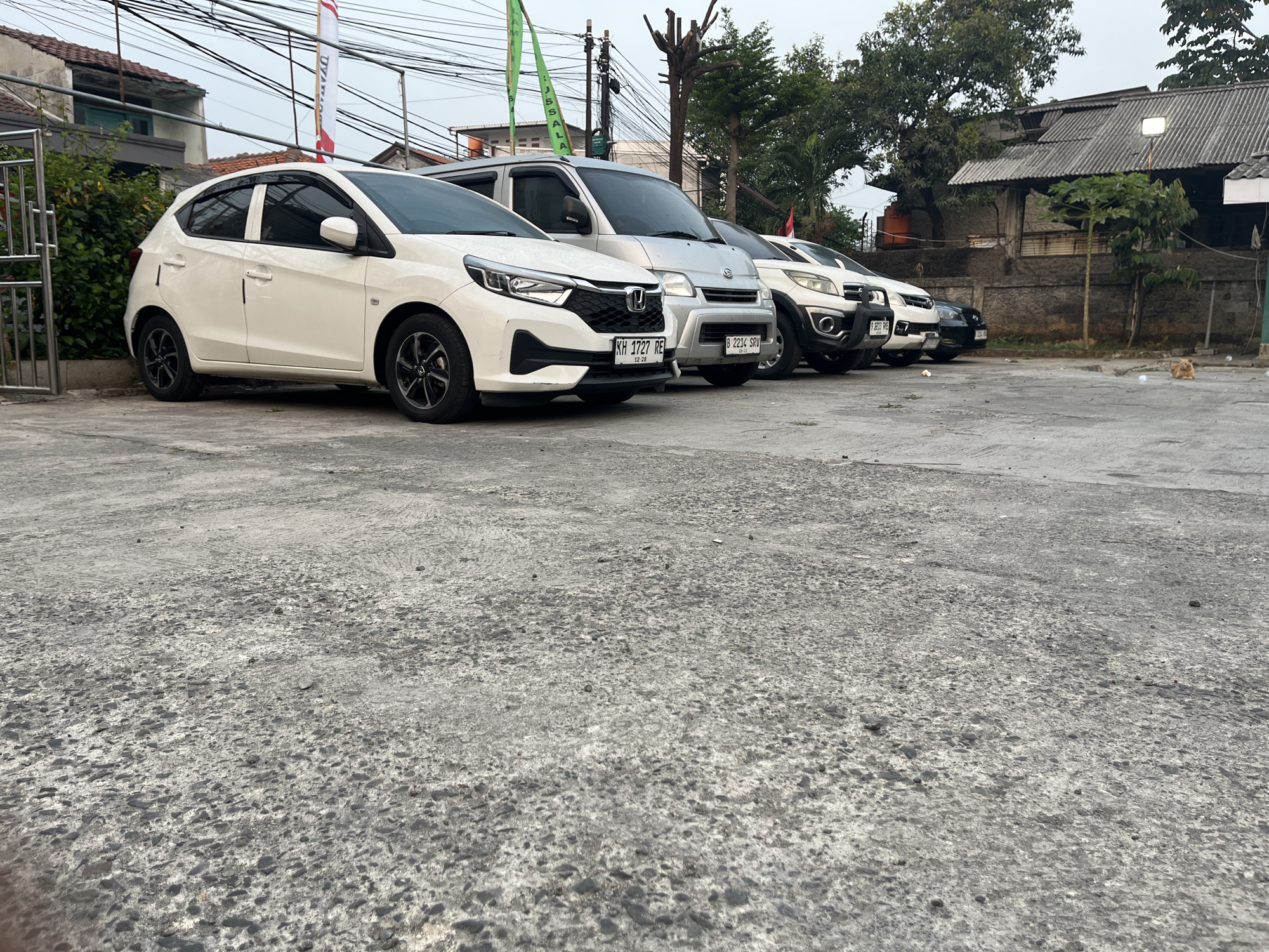 A line of parked cars is visible on a paved surface, likely a parking lot, in front of a row of buildings. The cars are mostly white and silver. The closest car is a white hatchback. Behind it are a silver minivan and other vehicles. In the background, there are buildings with corrugated roofs, trees, and electrical wires overhead. A small, orange cat is sitting on the pavement to the right of the frame. The scene appears to be located in a somewhat urban or suburban environment. The image was captured on October 7, 2023, at 17:09:00. While the specific location within East Jakarta, Indonesia, cannot be definitively determined from the image alone, the license plates on the vehicles (e.g., "KH 1727 RE" and "B 2234 SRV") suggest they are registered in Indonesia, and the architectural style of the buildings is consistent with that region.