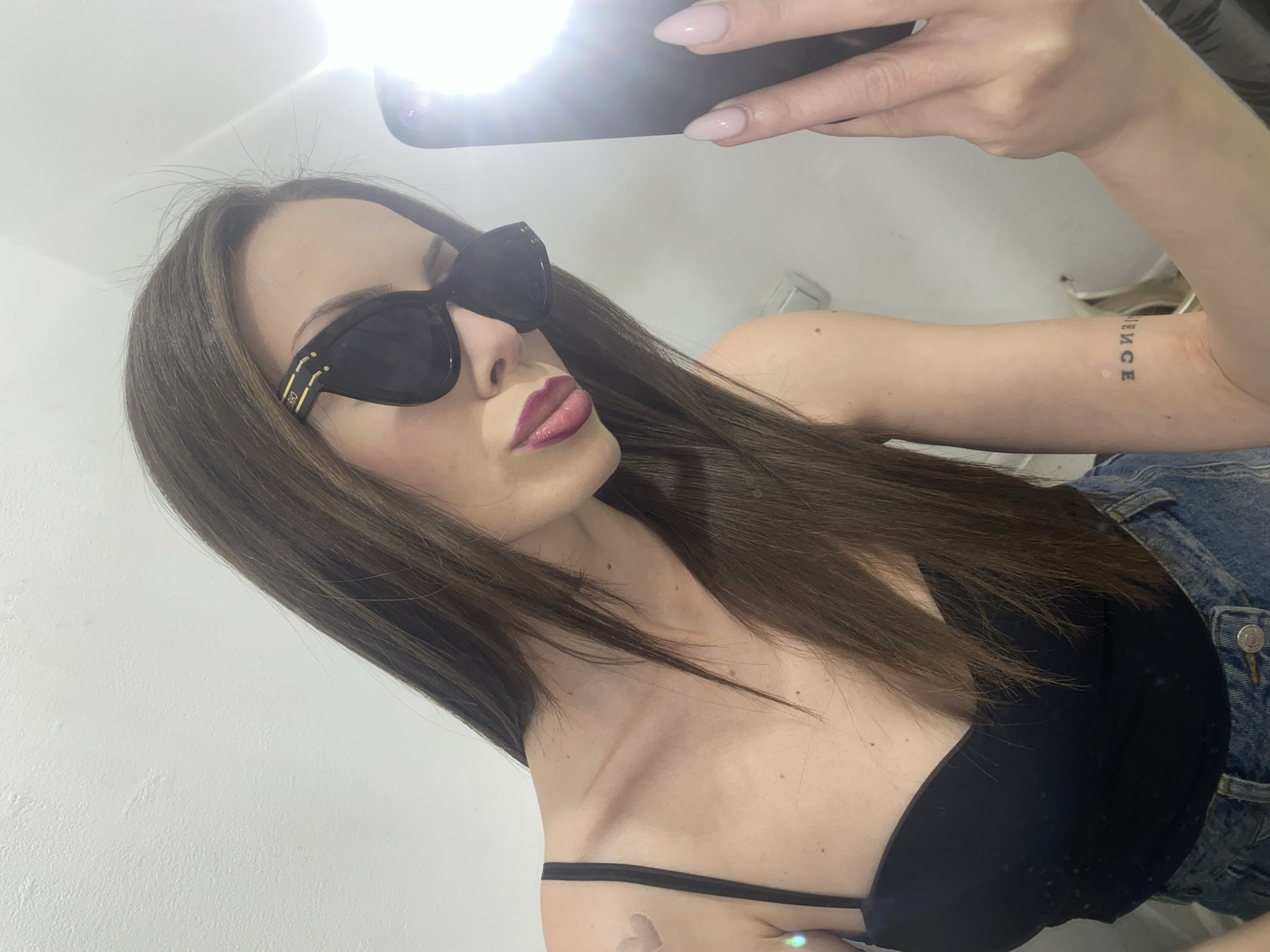 A young woman with long, dark brown hair is taking a selfie in a brightly lit room. She is wearing oversized black sunglasses, a black tank top, and denim shorts. She has on glossy pink lipstick and her nails are painted a light pink. She has a small heart tattoo on her left shoulder and a word tattoo on her right forearm. The background is a plain, light-colored wall. The lighting suggests an indoor setting, possibly a bathroom or dressing room, with a bright flash from her phone illuminating the scene.