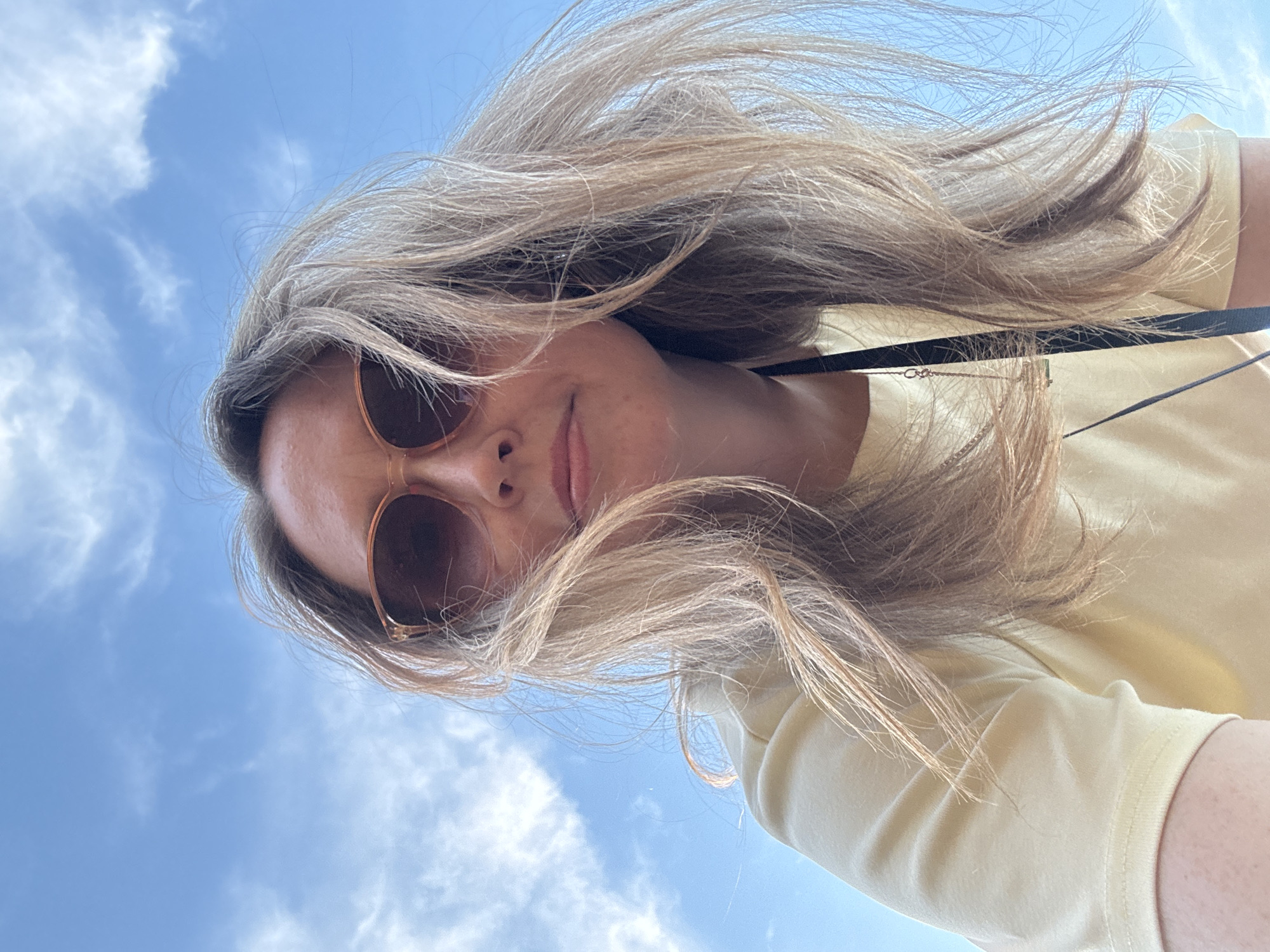 A woman is taking a selfie outdoors, looking up towards the camera. She has blonde hair, which is blowing in the wind, and is wearing sunglasses and a light yellow t-shirt. The sky is a clear blue with some white clouds. The image appears to be taken on a sunny day. The woman's face is partially obscured by her hair and sunglasses, but she appears to be smiling. There is no visible text in the image. The location context is Chaikovsky, Russia.