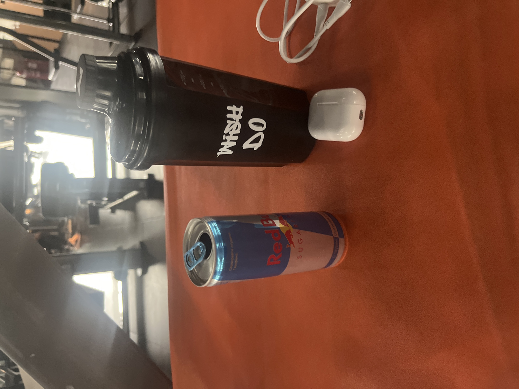 The scene is a gym in Sofia, Bulgaria. An open can of Red Bull, a black shaker bottle with the phrase "Wish Do" printed on it, a white AirPods case, and white earphones sit on an orange bench. Weight training equipment is visible in the background.