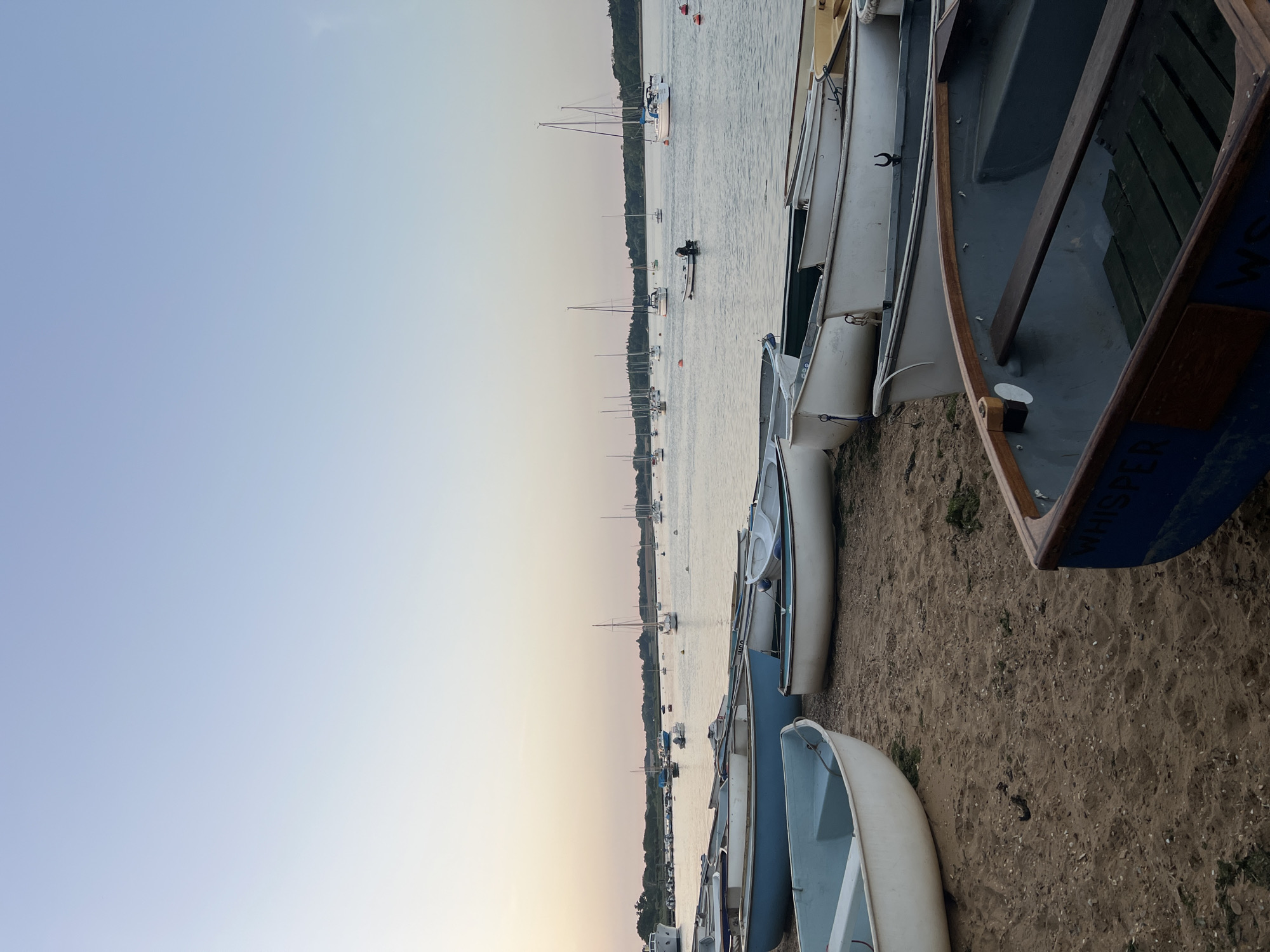 The image shows a row of small boats on a sandy beach. The boats are various shades of blue and white. In the background is a body of water with sailboats and trees on the far shore. The sky is a pale blue with hints of pink and yellow near the horizon, suggesting either sunrise or sunset. There is a person on a paddleboard in the water. The location is likely near a body of water in East Suffolk, United Kingdom, based on the vegetation and boat types.
