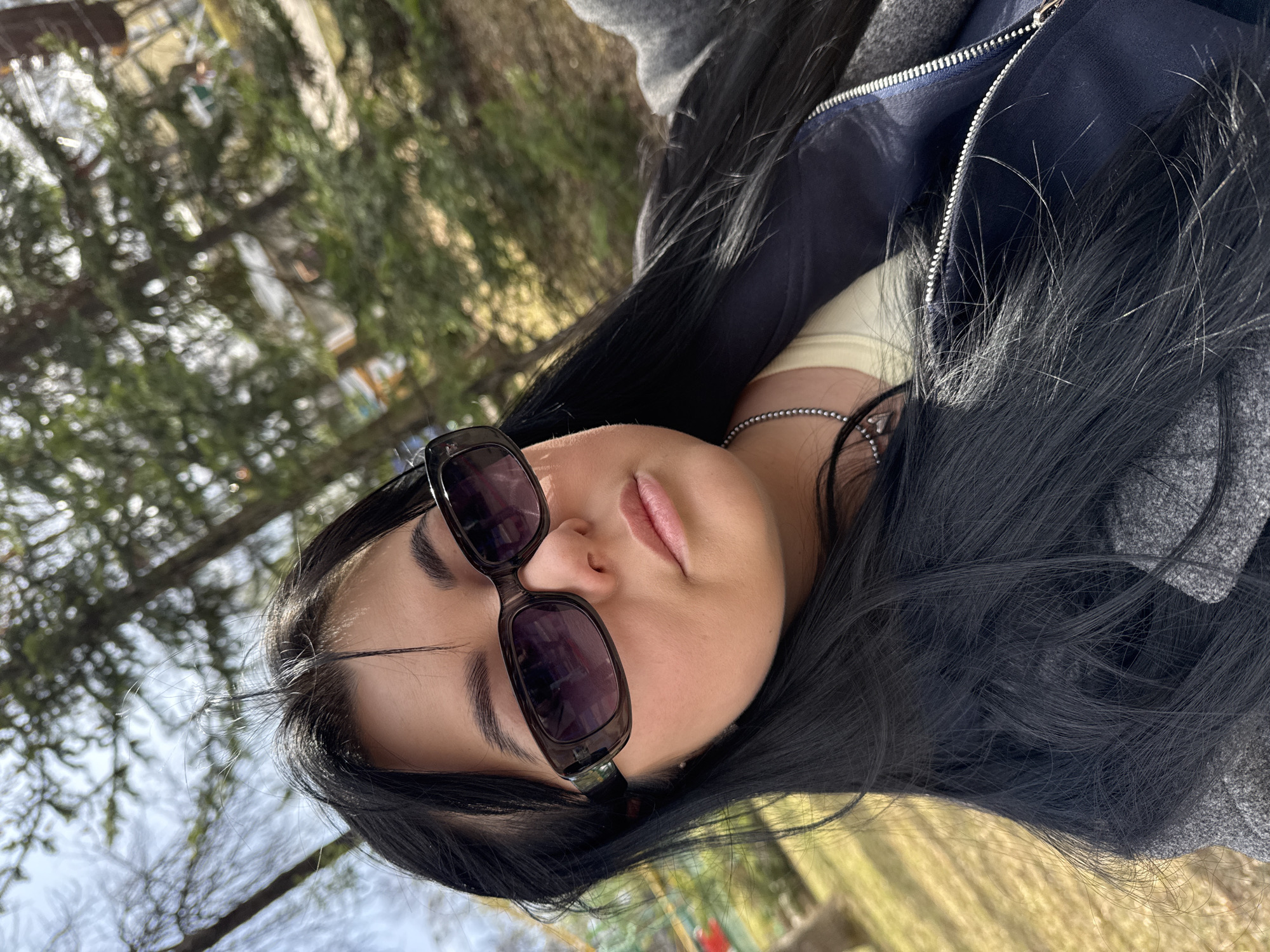A young woman with long, black hair and sunglasses is lying down outdoors. She is wearing a light-colored shirt, a chain necklace, and a gray jacket with a navy blue interior visible near the zipper. The background includes trees and grass, suggesting a park or green area. The location is Perm, Russia.