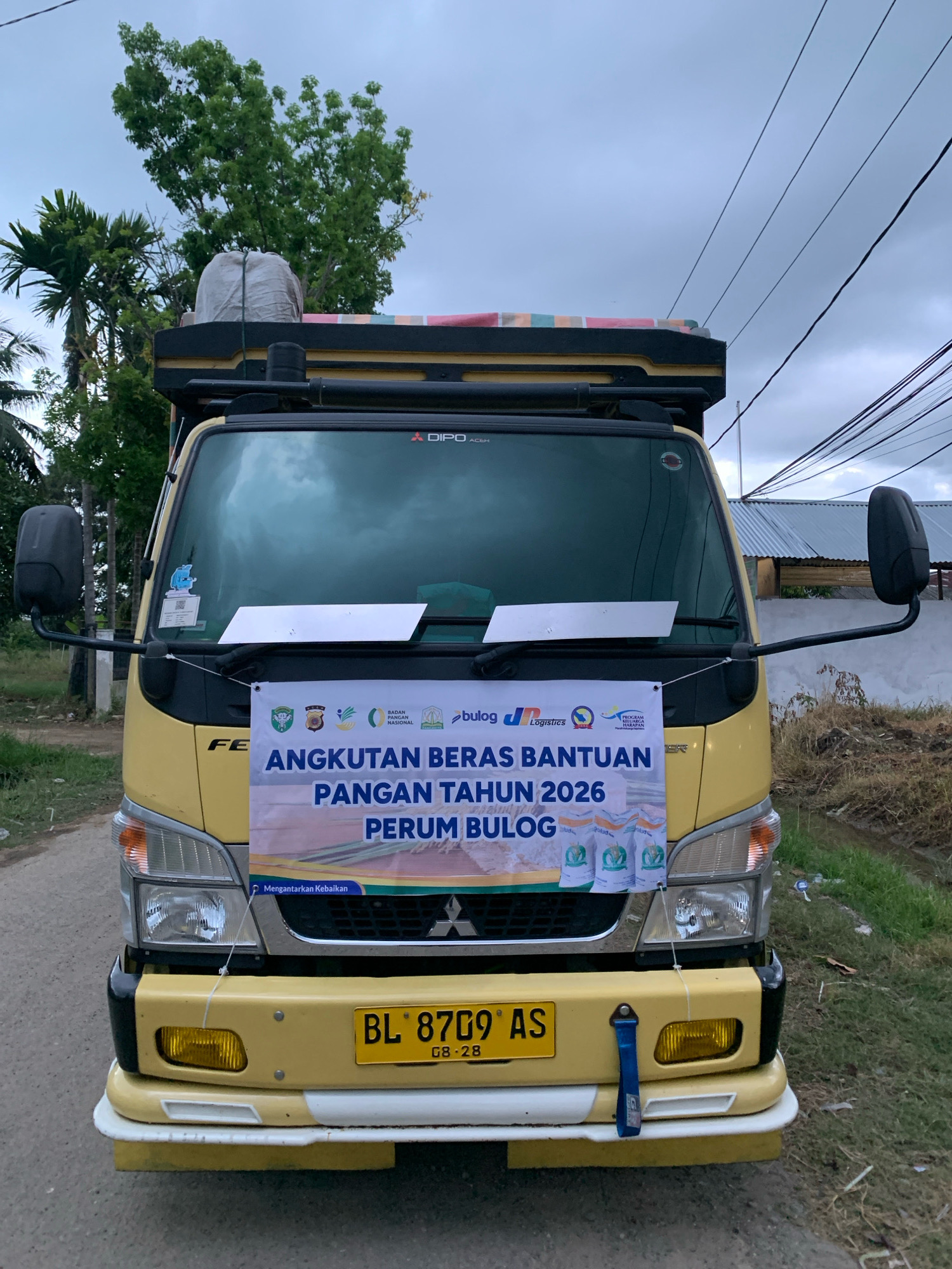 A yellow truck is parked on a paved road in Pantegurah, Indonesia, under an overcast sky. The truck is adorned with a banner that reads "ANGKUTAN BERAS BANTUAN PANGAN TAHUN 2026 PERUM BULOG," indicating it is carrying rice aid for the year 2026 from Perum Bulog, an Indonesian state-owned logistics company. The banner also features logos of several organizations, including Badan Pangan Nasional and Logistrcs. The truck's license plate reads "BL 8709 AS" with "08-28" below it. To the left of the truck, palm trees and other greenery are visible. The background shows a building with a corrugated metal roof and some dry vegetation. The overall scene depicts a logistics operation involving food distribution.