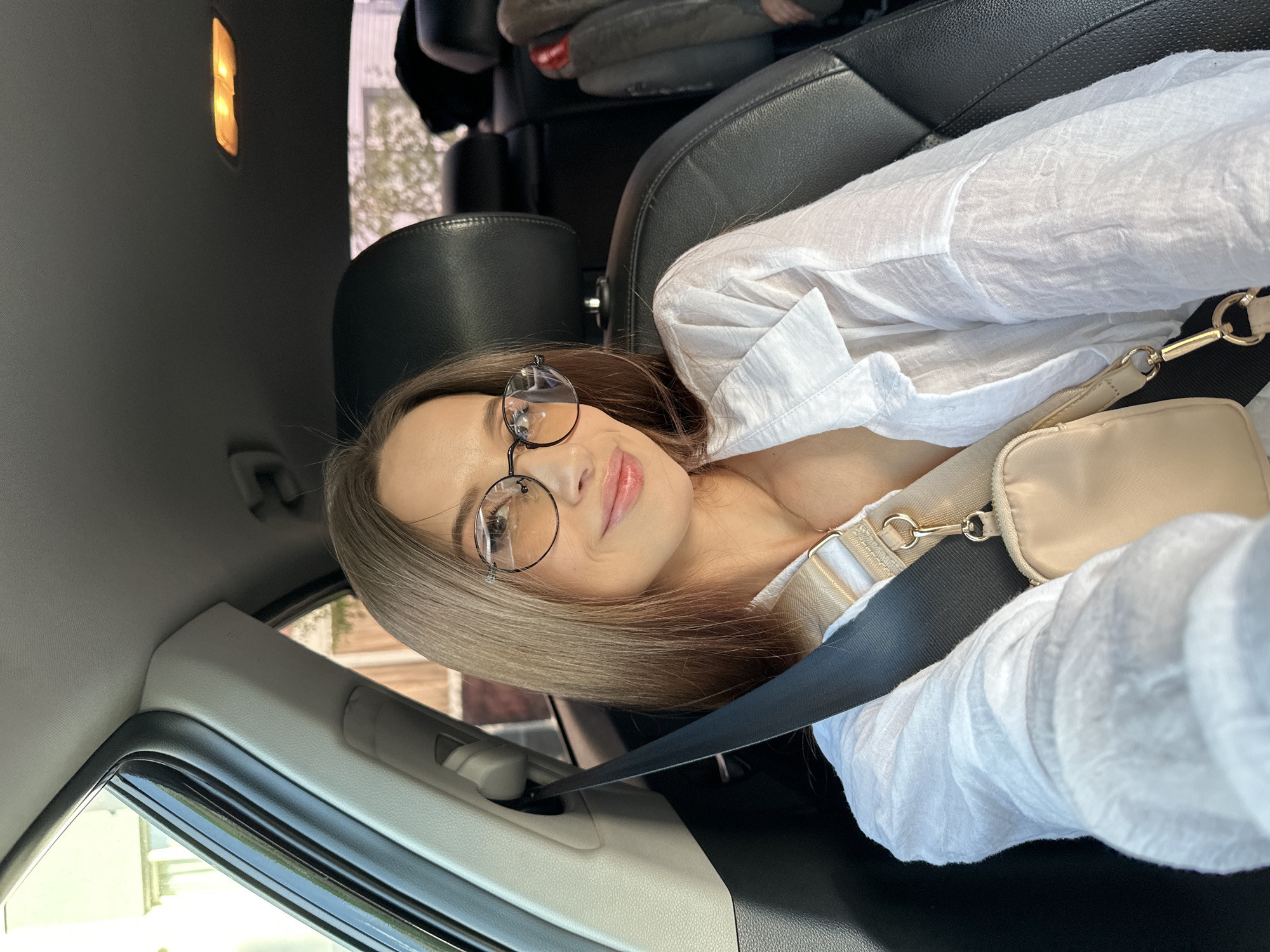 A young woman with light brown hair and glasses sits in the driver's seat of a car, wearing a white shirt and a seatbelt. She is angled towards the camera and smiling. The interior of the car is visible, including the dashboard, window, and overhead light. Outside the car window, buildings and trees can be seen, giving a general sense of being in a city. The city location in Yekaterinburg, Russia, cannot be determined with certainty.