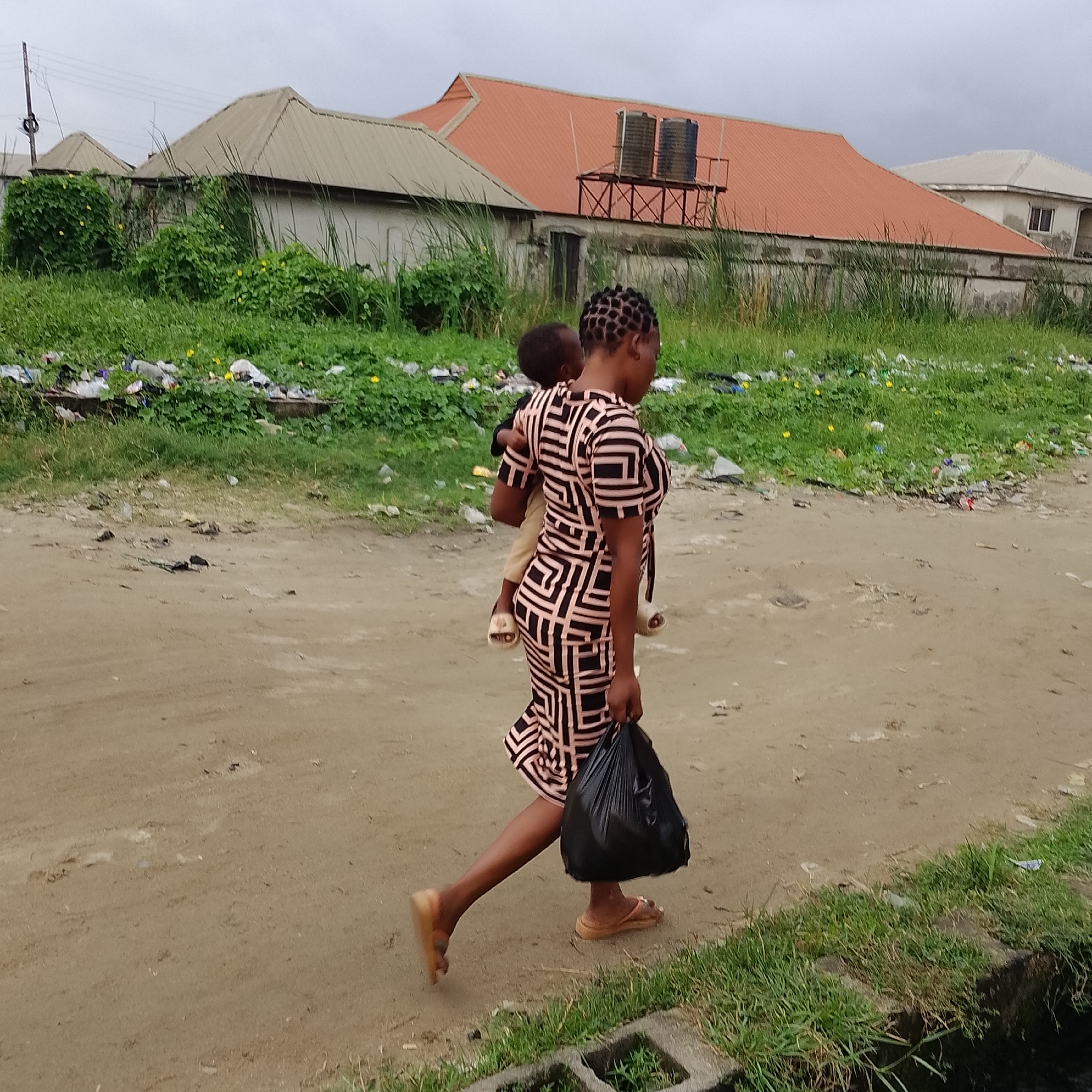 The image depicts a street scene in Ado, Nigeria, on an overcast day. A woman is walking away from the camera, carrying a young child on her back. She is wearing a black and pink geometric-patterned dress and appears to be wearing flip-flops. In her right hand, she is carrying a black plastic bag. The ground is a dirt road with patches of grass along the side. In the background, there are residential buildings with corrugated metal roofs, some with visible water tanks on top. There is also evidence of litter and overgrowth on the side of the road. A power line pole is visible in the far left background.
