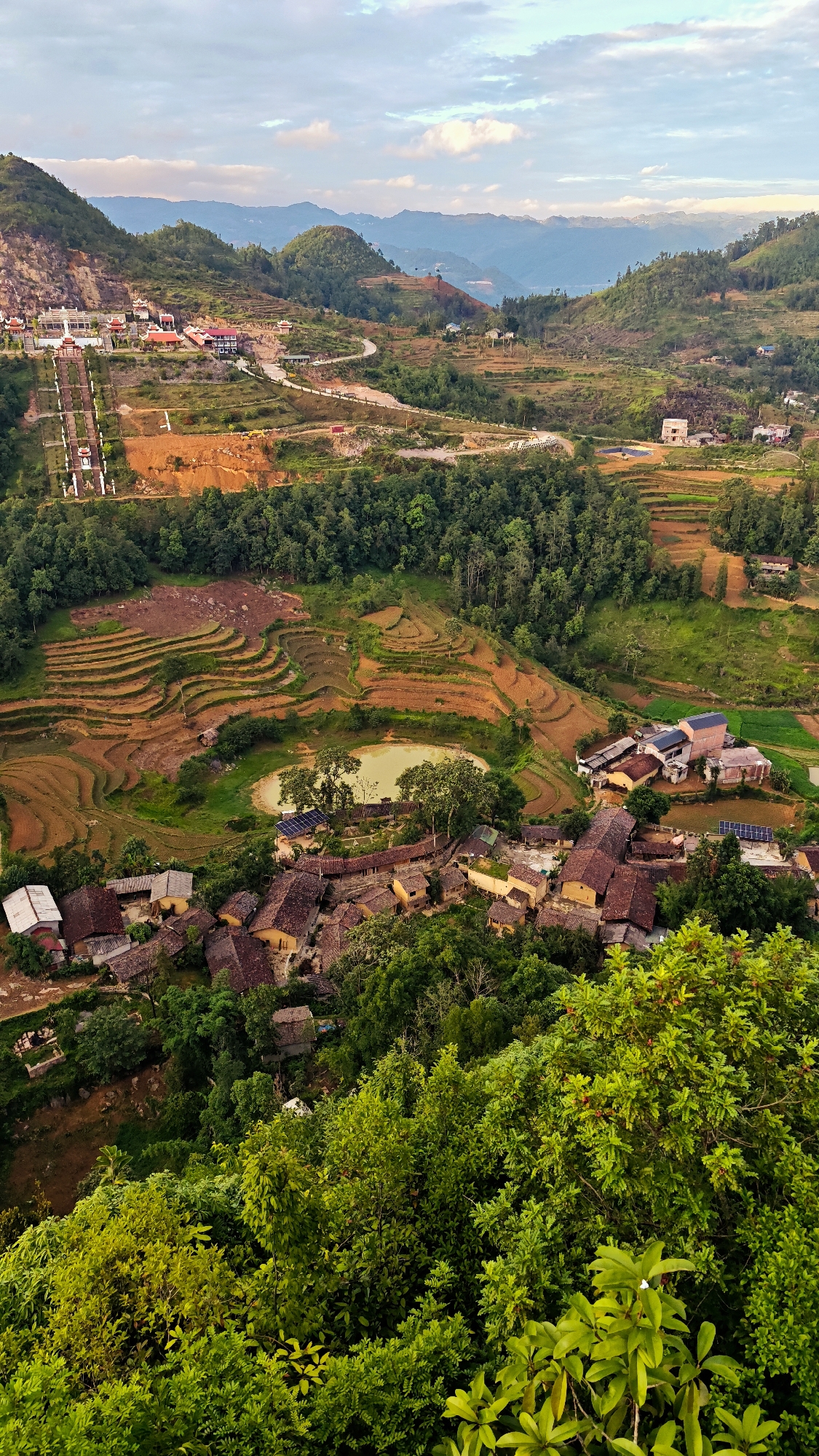 The image depicts a scenic overview of Lũng Cú Commune in Vietnam. In the foreground, lush green vegetation dominates the lower right, gradually transitioning to a village with houses featuring brown tile roofs nestled among the trees. Beyond the village, terraced fields are visible, showing a mix of cultivated plots and bare earth. A small body of water, surrounded by trees, is present near the village. In the mid-ground, a combination of dense forest and open areas with more scattered vegetation is seen. Towards the upper left, a complex of buildings with red-tiled roofs and a series of steps leading up a hillside are apparent. Mountains are visible in the background, under a cloudy sky. A road meanders through the landscape, connecting different areas.