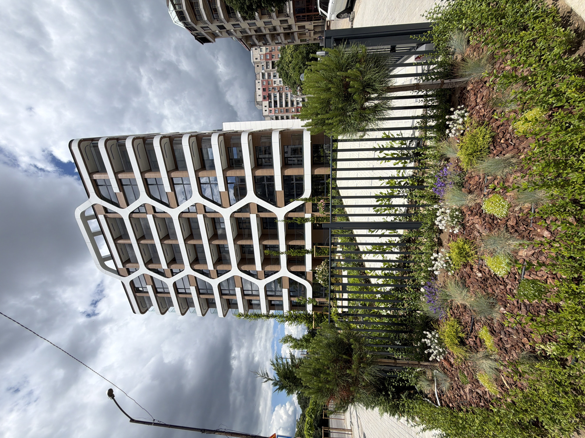 This is a high-angle, full shot of a modern, multi-story building in Sofia, Bulgaria, under a cloudy sky. The building has a distinctive honeycomb-like facade with white geometric patterns overlaying brown and glass panels. Balconies with glass railings are visible on each floor. In the foreground, there is a neatly landscaped garden with bark mulch, various low-lying plants, and small trees, enclosed by a black metal fence. A streetlight stands to the left of the building. Other buildings and trees are visible in the background, including a taller, older apartment building with red accents. The street and sidewalk are paved with light-colored tiles. No people are clearly visible.