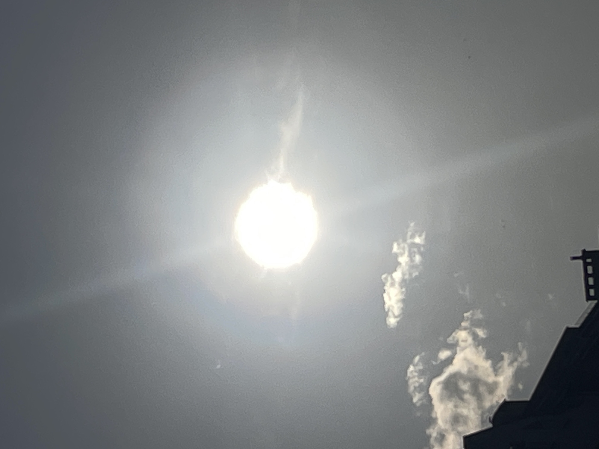 A bright sun is centered in a hazy, gray sky, casting a lens flare across the image. Several wispy clouds are visible in the lower half of the frame, and the silhouette of a building with architectural details can be seen in the bottom left corner. The scene appears to be outdoors, with no people or identifiable actions occurring.