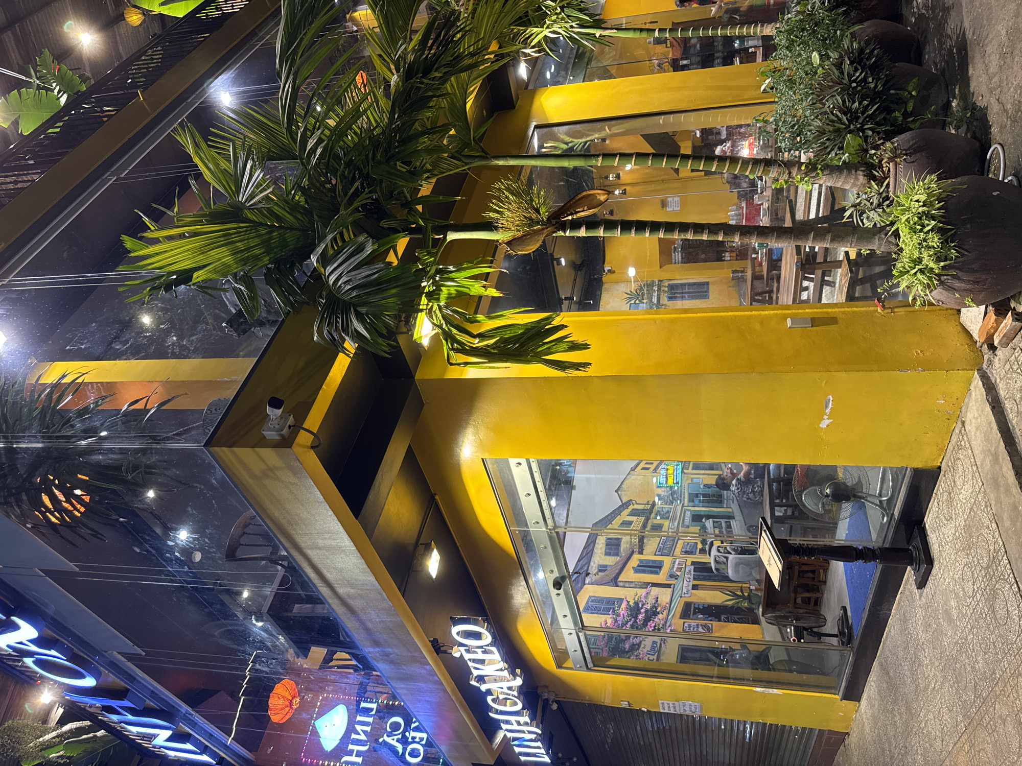 A vibrant yellow building with large glass windows is illuminated at night in Nha Trang Ward, Vietnam. Palm trees with lush green fronds are strategically placed both inside and outside the building, some in large dark planters. Inside, through the glass, a restaurant or cafe setting is visible, with tables and chairs arranged, and a few people can be seen. A ceiling fan is also visible inside, as is a floor fan near the window. The exterior features neon signs with blue and white lettering, one of which appears to read "LINH CAO" and another with what seems to be Vietnamese characters and a graphic of a lantern. The overall scene is colorful and inviting, suggesting a lively evening atmosphere. The building's architecture is modern, with geometric angles and reflective surfaces. The time of day is clearly night, with the building's lights and the neon signs creating a warm glow against the dark surroundings.