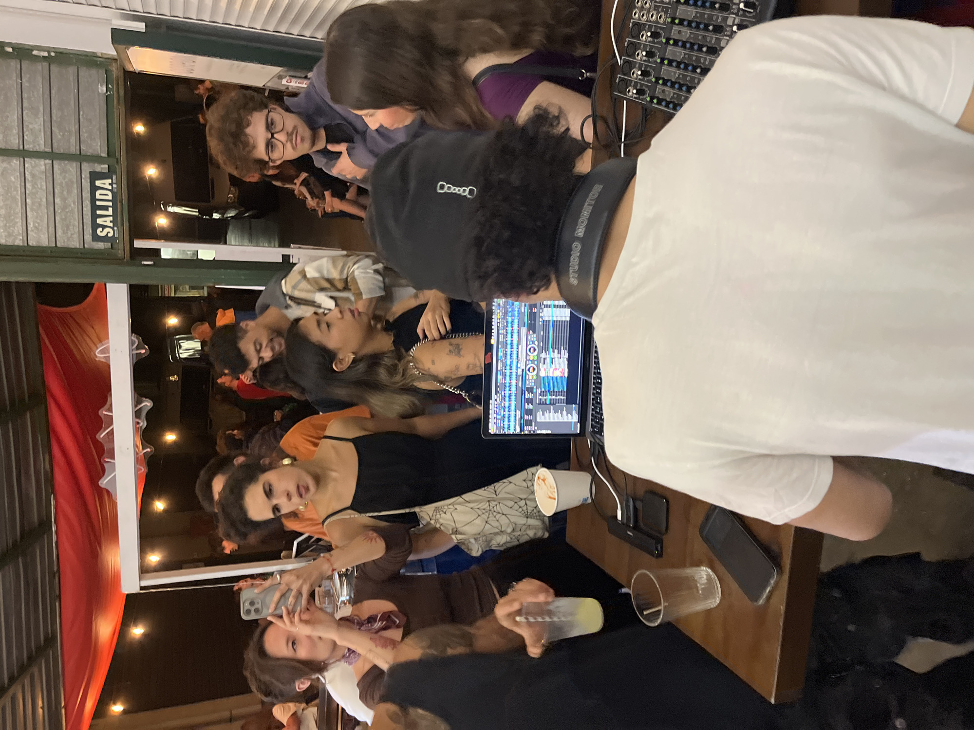 The image captures an indoor scene in Barcelona, Spain. A group of people are gathered around a DJ who is working on a laptop connected to a mixing board. The DJ is wearing a black cap and headphones with the text "STUDIO MONITOR" visible. A sign with the word "SALIDA" (Exit in Spanish) is present. People are holding drinks, looking at phones, and interacting with each other and the DJ. The setting appears to be a casual gathering or party, possibly in a bar or club.