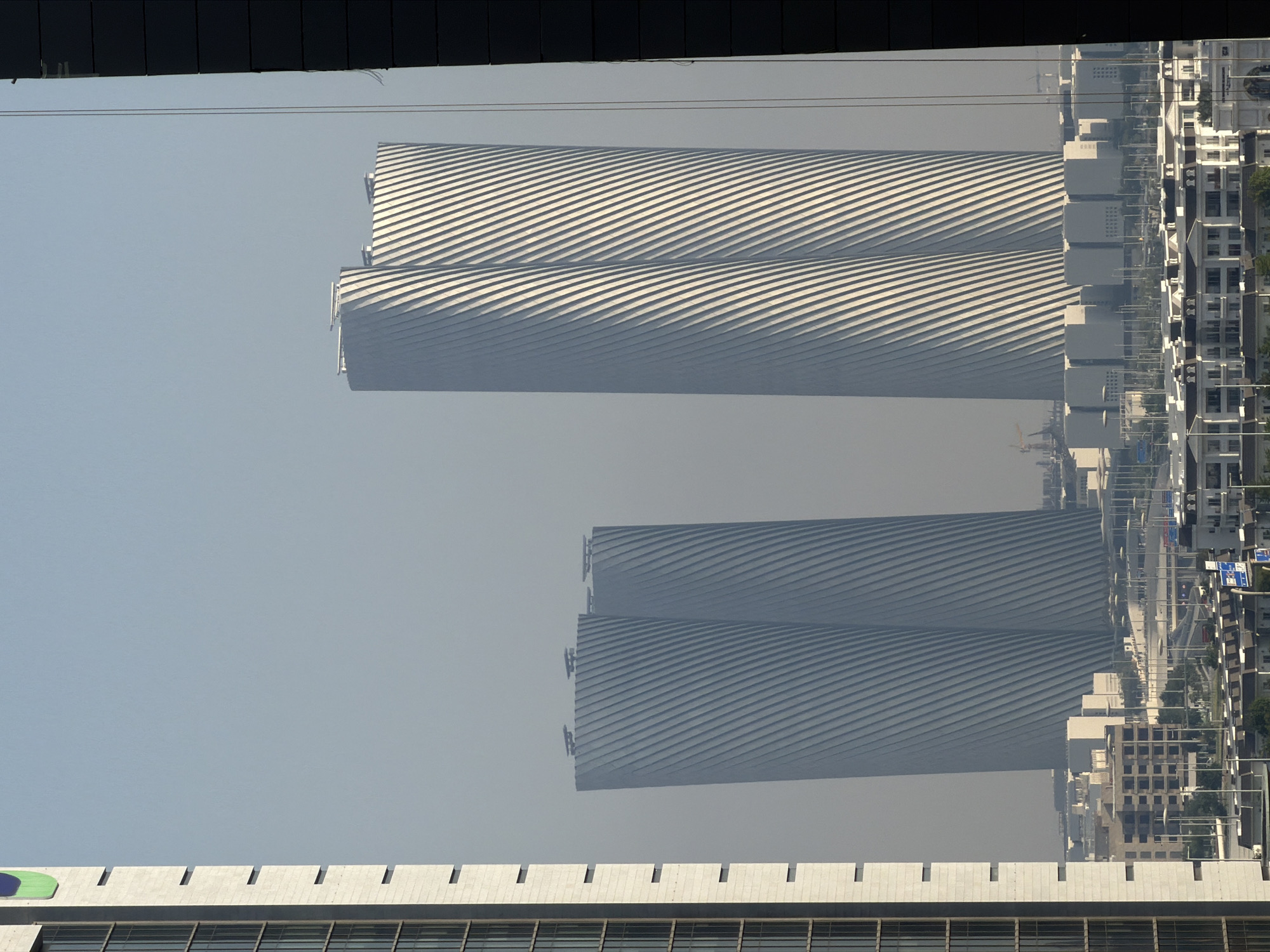 The image shows a city skyline with a hazy sky. In the foreground, on the left, a tall, light-colored building with a green and blue circular logo is visible. To the right of this, in the midground, are two large, white, cylindrical skyscrapers with a unique twisted facade characterized by diagonal lines. Further in the background, more buildings of varying sizes and architectural styles are visible, along with roads and streetlights. The overall impression is one of modern urban development under a slightly overcast or dusty atmosphere.