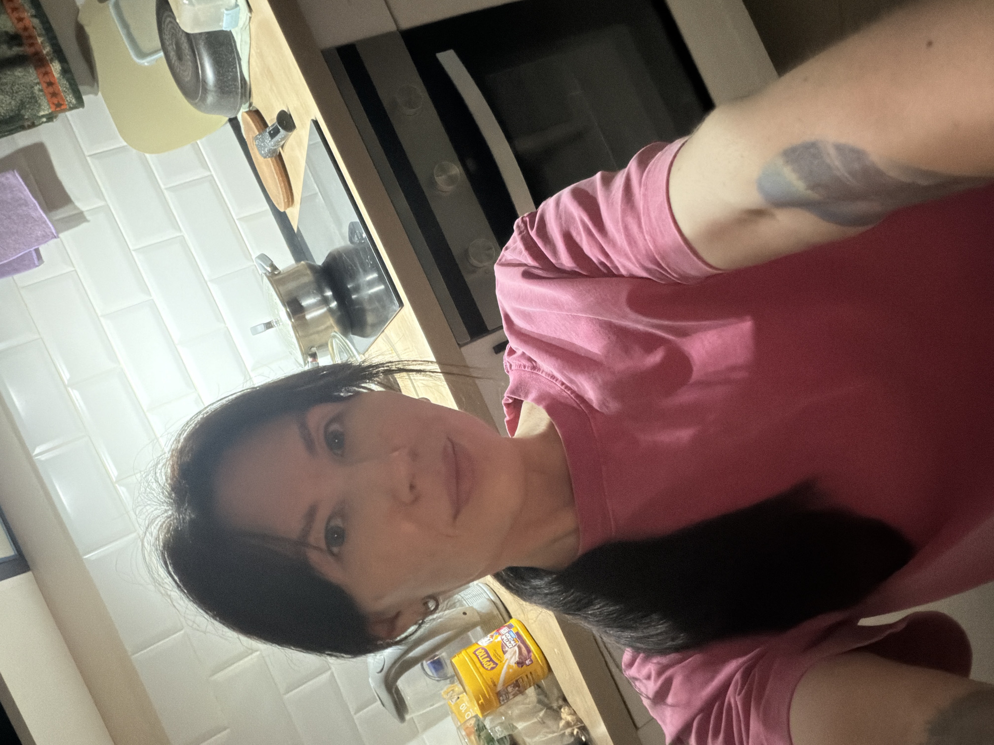 A selfie shows a woman with dark hair and a pink shirt in a kitchen in Perm, Russia. She has a tattoo on her left arm. The kitchen features white subway tile, wooden countertops, and a built-in oven. There's a sink with a faucet, a pot on the stove, and various kitchen items on the counter, including a package of "Vollage" crackers.