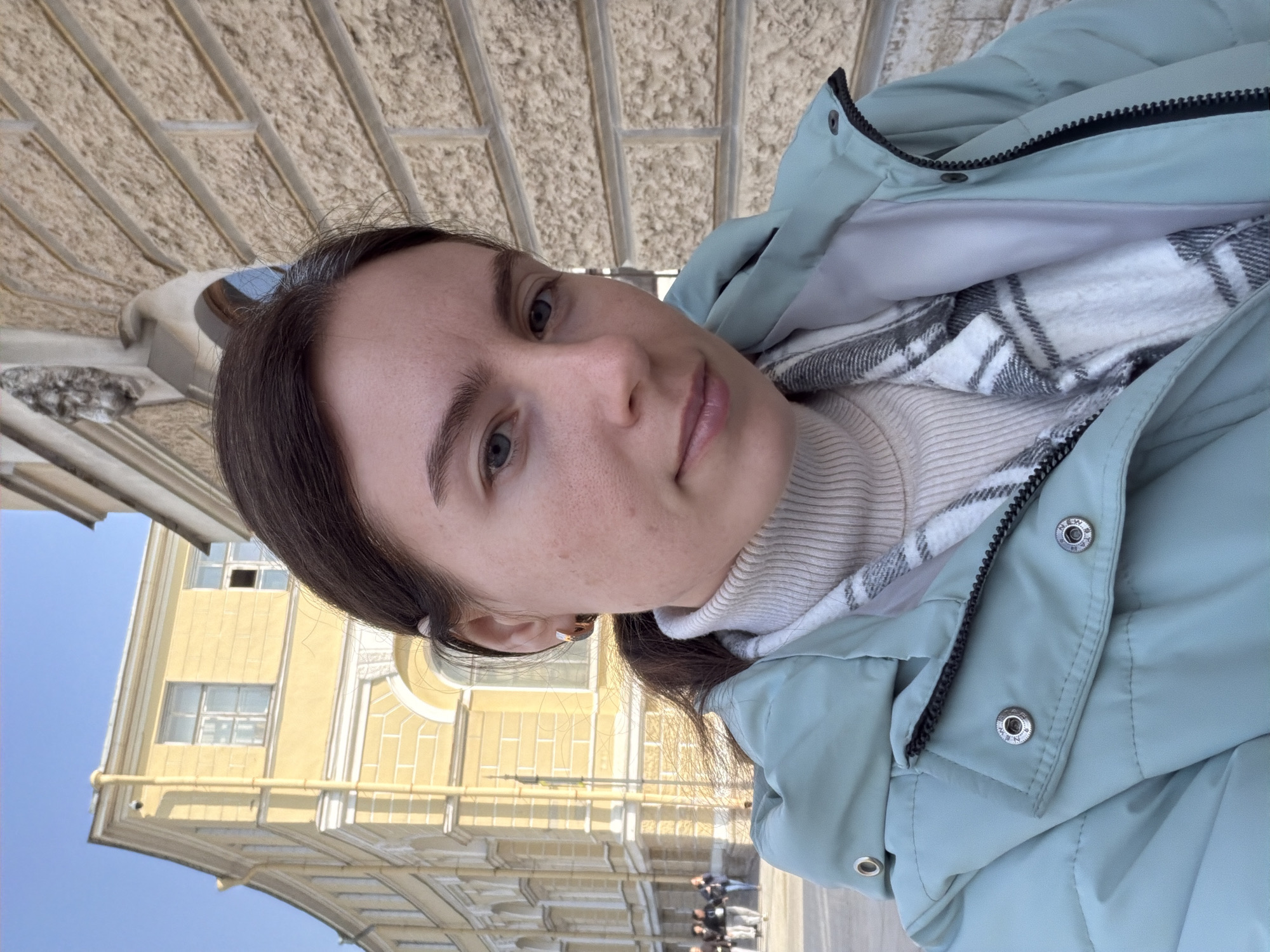A woman is standing outside in Saint Petersburg, Russia. She is wearing a light blue jacket over a white turtleneck and a plaid shirt. A yellow building with ornate windows is behind her. Other people can be seen in the background. The shot is angled, suggesting the person holding the camera is shorter or the camera is held at an angle. The lighting indicates it is daytime.