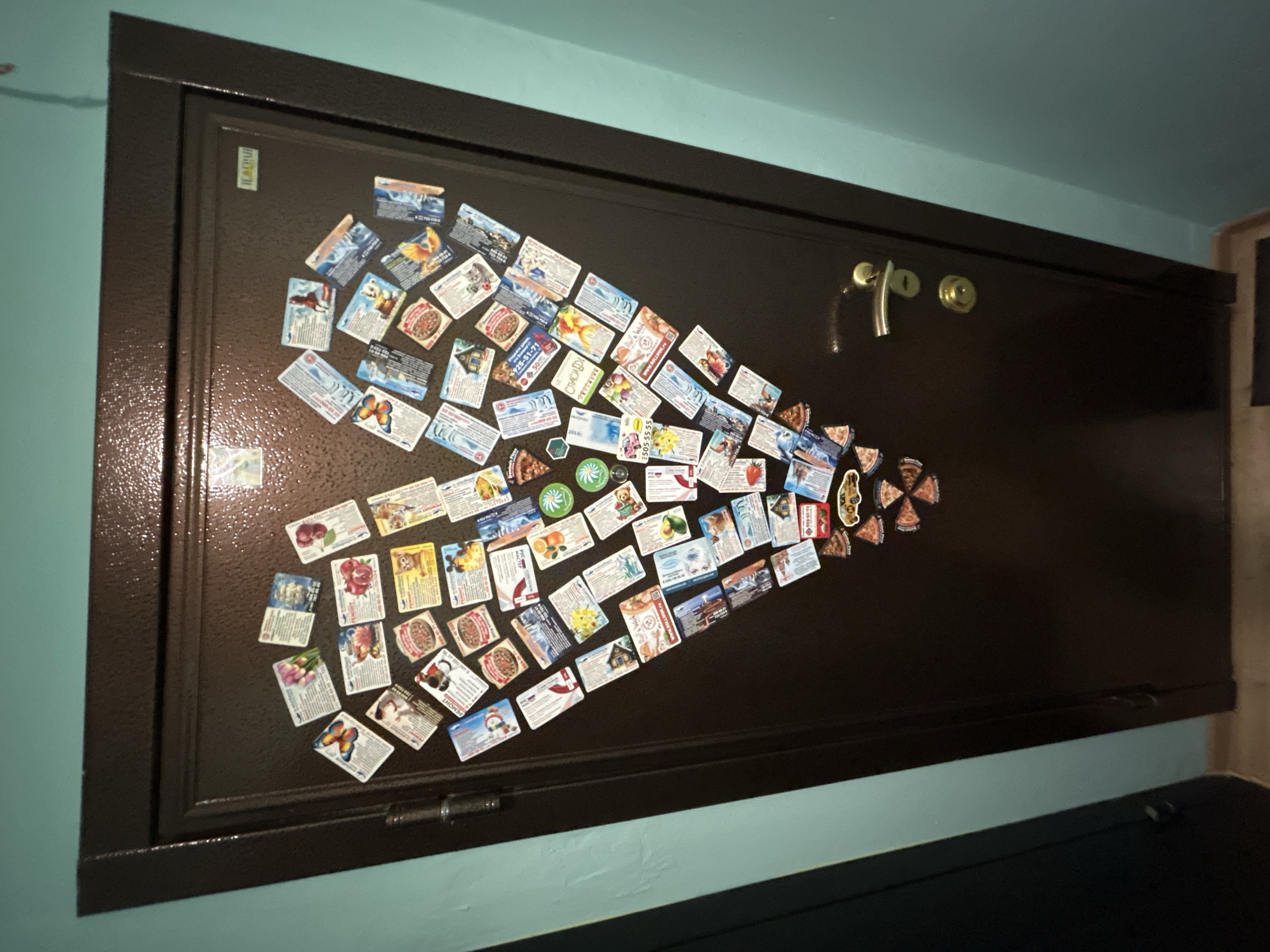 A wide shot depicts a brown metal door, likely a steel security door, mounted on a wall painted in light blue. The door is decorated with numerous small rectangular cards or magnets arranged in a rough heart shape. The door has a locking mechanism with a handle and a keyhole. The overall image is slightly out of focus, and the scene appears to be indoors. There is no explicit evidence within the image to definitively confirm the location is Moscow, Russia.