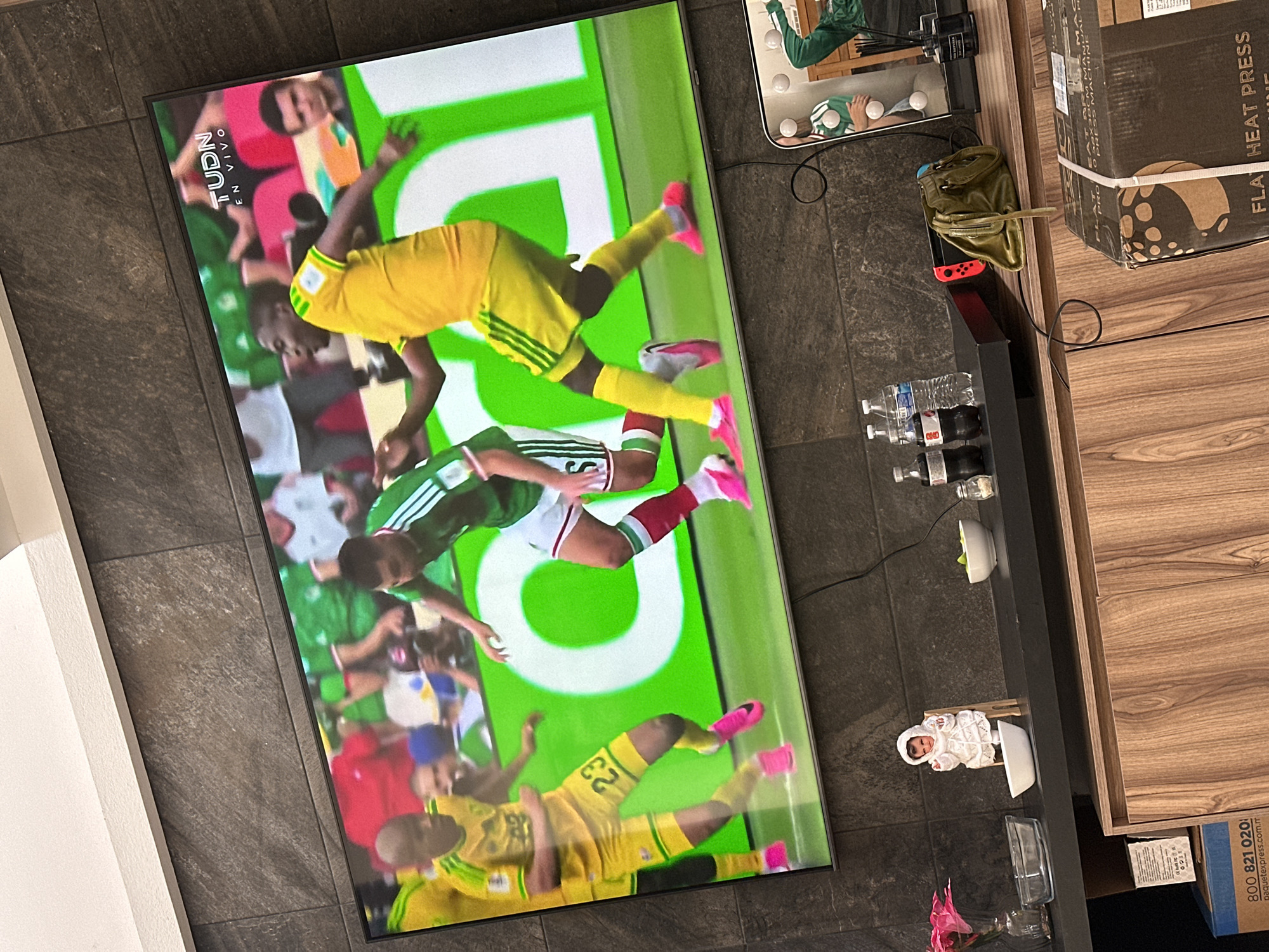 In a living room setting in San Bernardino Tlaxcalancingo, Mexico, a large television screen displays a live broadcast of a soccer match. Two male athletes are prominently featured, one in a yellow jersey with the number 23, and another in a green and white jersey. The background on the screen shows a crowd and stadium signage. In the foreground, on a black shelf beneath the television, several items are visible: a decorative figurine of a baby in white, two bottles of soda, a bottle of water, a small white bowl with green food, a clear glass bowl, and a pink flower arrangement in a vase. To the right of these items, a Nintendo Switch console is visible, with a green handbag placed on top of it. A vanity mirror with lights is also present in the background on the right side of the shelf. Below the shelf, a wooden cabinet is partially visible. A large cardboard box with text partially obscured is in the lower right corner of the image.