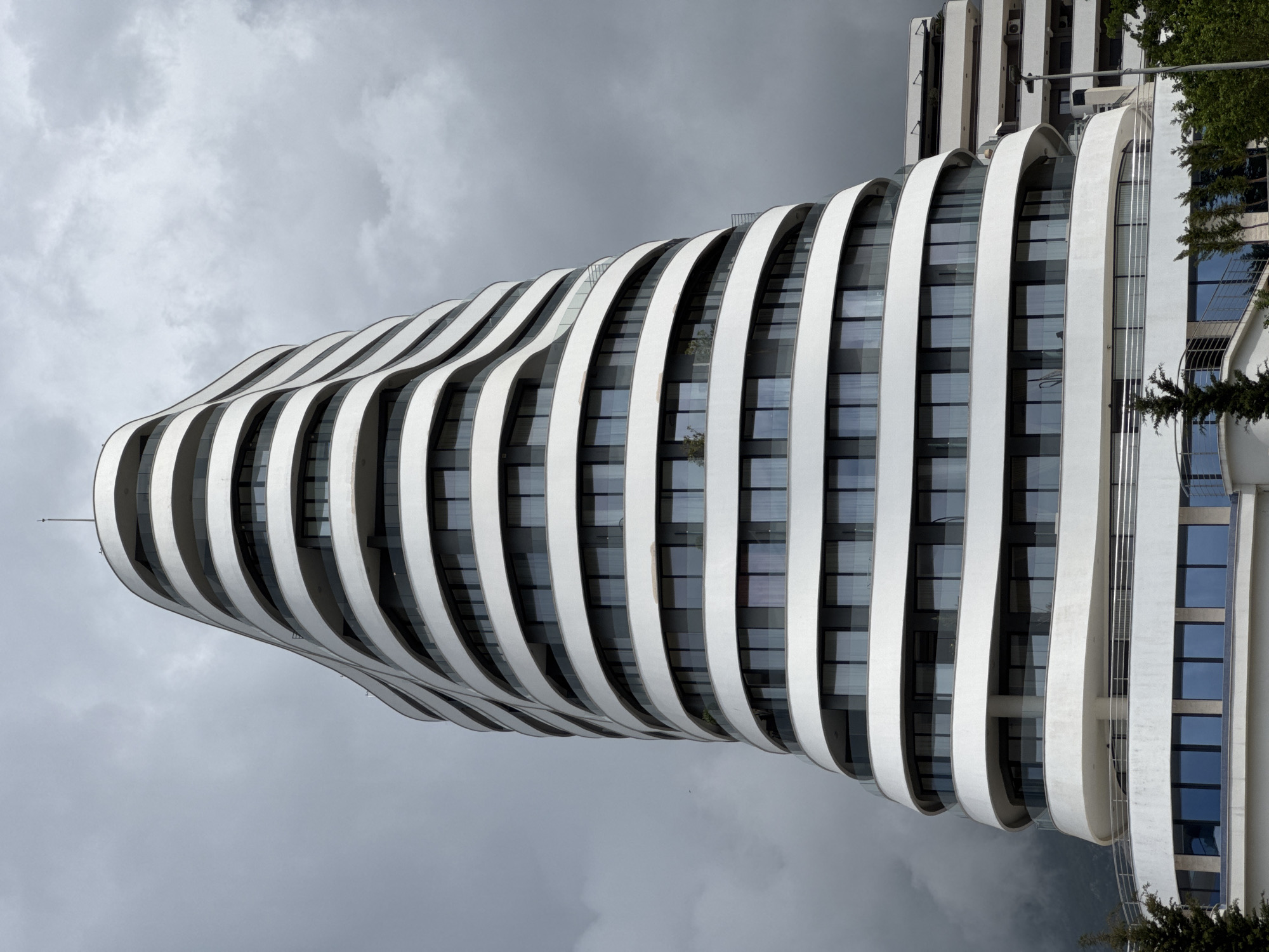 An eye-level, slightly-angled photograph depicts a contemporary white high-rise building in Boreti, Montenegro against an overcast sky. The building features a distinctive curved architectural design with alternating horizontal white bands and glass windows. The white bands appear as continuous curvilinear balconies or ledges. A secondary building is visible in the upper right portion of the photo. The photo is taken from a position slightly below the building, and the image has been rotated 90 degrees counter-clockwise.