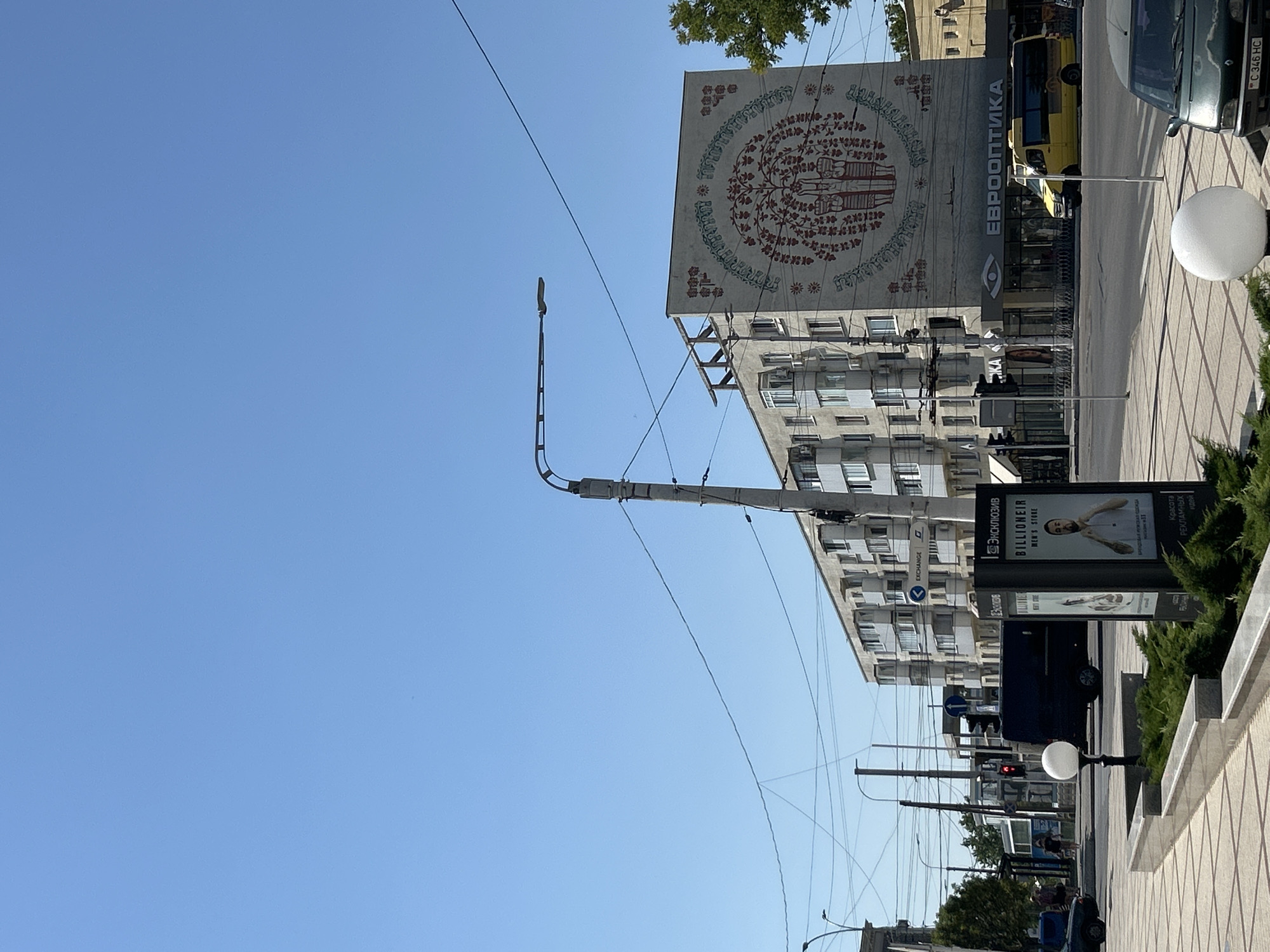 The image depicts a street scene in Tiraspol, Moldova, on a clear, sunny day. In the background, a multi-story apartment building with a large, stylized mural dominates the view. The mural features a tree-like motif with figures and decorative elements in red and green. Below the mural, a store with the name "ЕВРООПТИКА" (Eurooptika) is visible, indicated by an eye logo and text. A yellow bus is parked in front of the store.
In the foreground, a sidewalk with a patterned pavement runs along the street. Several streetlights and overhead wires are present. A dark-colored van is stopped at a traffic light, which is red. To the right of the van, a billboard displays an advertisement for a men's clothing store named "BILLIONEIR." The billboard features a man in a white shirt. A few people are visible walking on the sidewalk in the middle ground. The environment appears to be urban, with a mix of residential and commercial buildings. A green tree is also visible on the right side of the image. The license plate of a car in the lower right corner reads "C 346 HC."