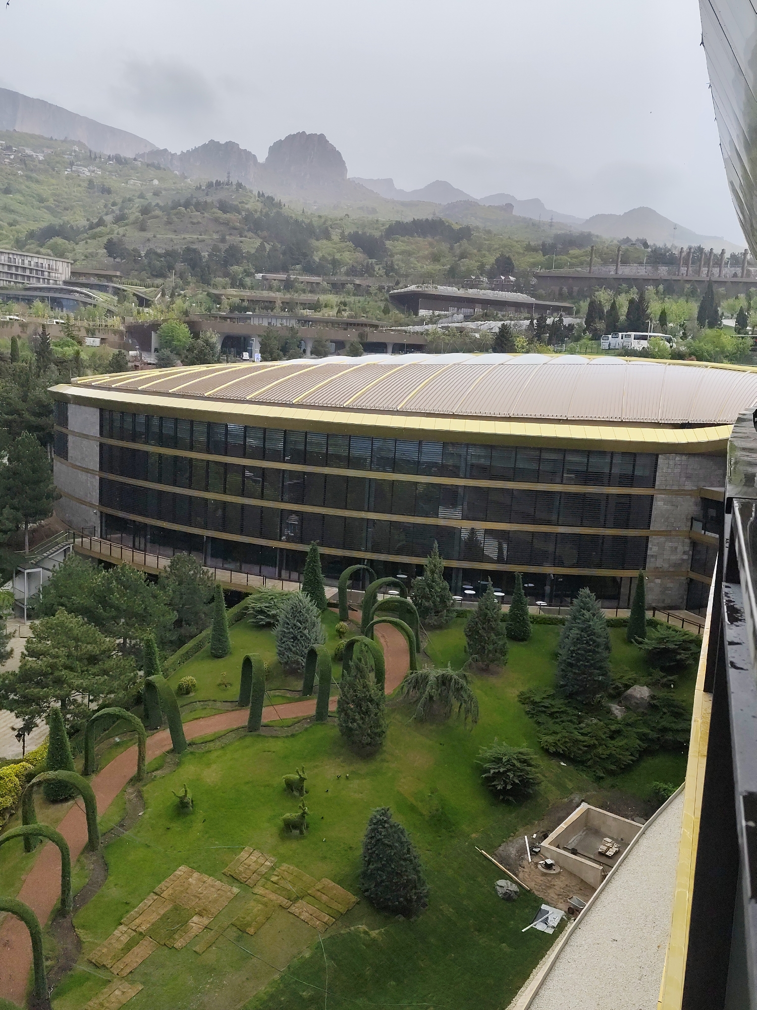This is a high-angle view of a building and garden in Симеизский поселковый совет, Ukraine, on a cloudy day. The building is modern, with a gold-colored roof and multiple tiers of windows. A well-manicured garden stretches out below, featuring topiary arches, winding pathways, evergreen trees, and decorative shrubbery. At least one construction project is in progress in the garden. In the background, the hazy outline of the Crimean Mountains is visible. Other structures and vegetation fill the mid-ground. The perspective suggests the photo was taken from a higher point, possibly another building.