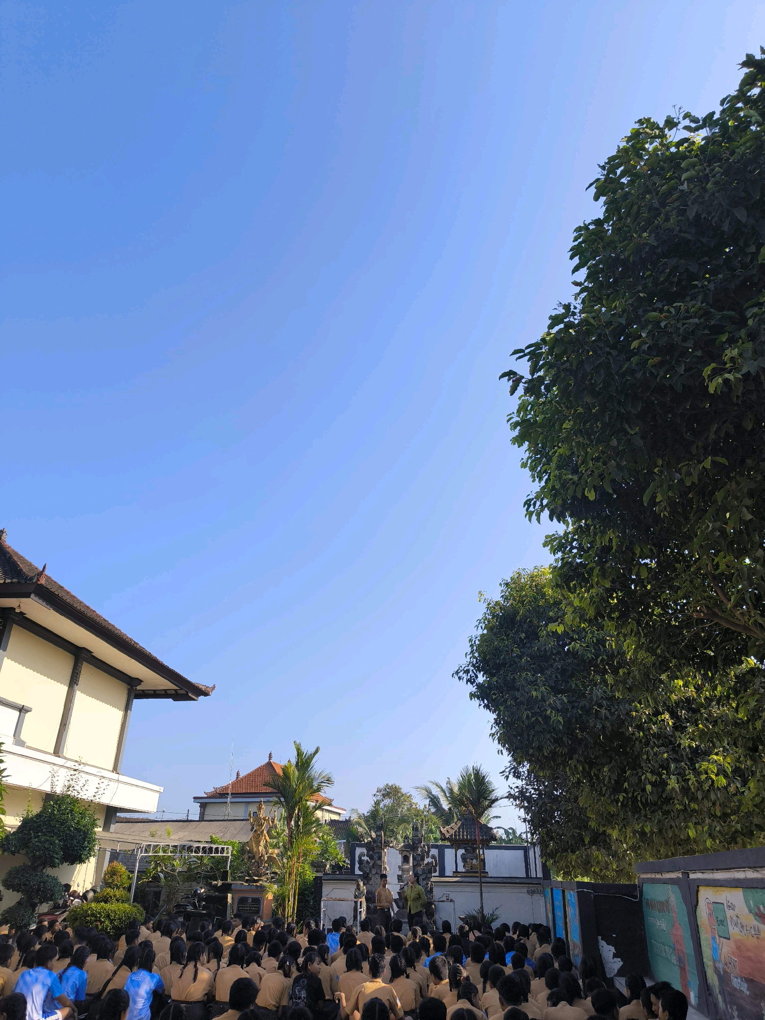 An outdoor scene captures a gathering of students in a courtyard, likely at a school, under a clear blue sky. The students, dressed in uniform khaki shirts and dark pants or skirts, are seated on the ground facing a raised area. In the background, a traditional Indonesian architectural style is visible, with a building featuring a dark, multi-tiered roof and cream-colored walls on the left. Adjacent to this building is a decorative gate, possibly leading to a temple or another significant structure. A statue of a deity or mythical figure stands prominently near the gate. Lush green trees and foliage surround the courtyard, with a large tree dominating the right side of the frame. Several individuals, presumably teachers or presenters, are standing in front of the seated students, facing them. The overall impression is one of an organized event or assembly taking place in a culturally rich environment. The location is specified as Tabanan, Indonesia.