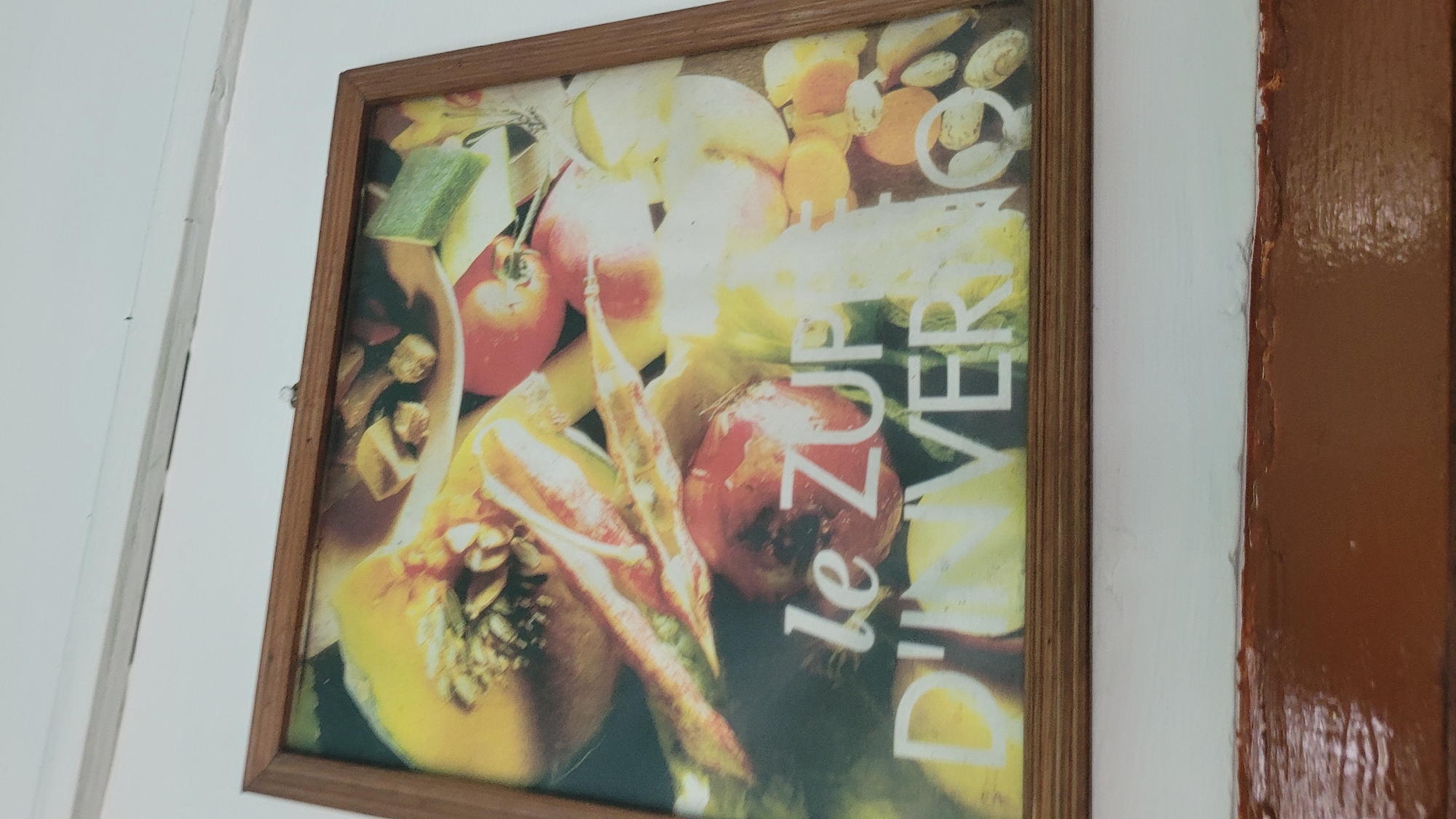 A framed photograph depicts a still life of vegetables and fruits, primarily in warm tones of yellow, orange, and red. In the foreground, large white text reads "le ZUPPE D'INVERNO". The photograph is hanging on a white wall above a dark brown wooden ledge. This image was taken in Vashisht, India.