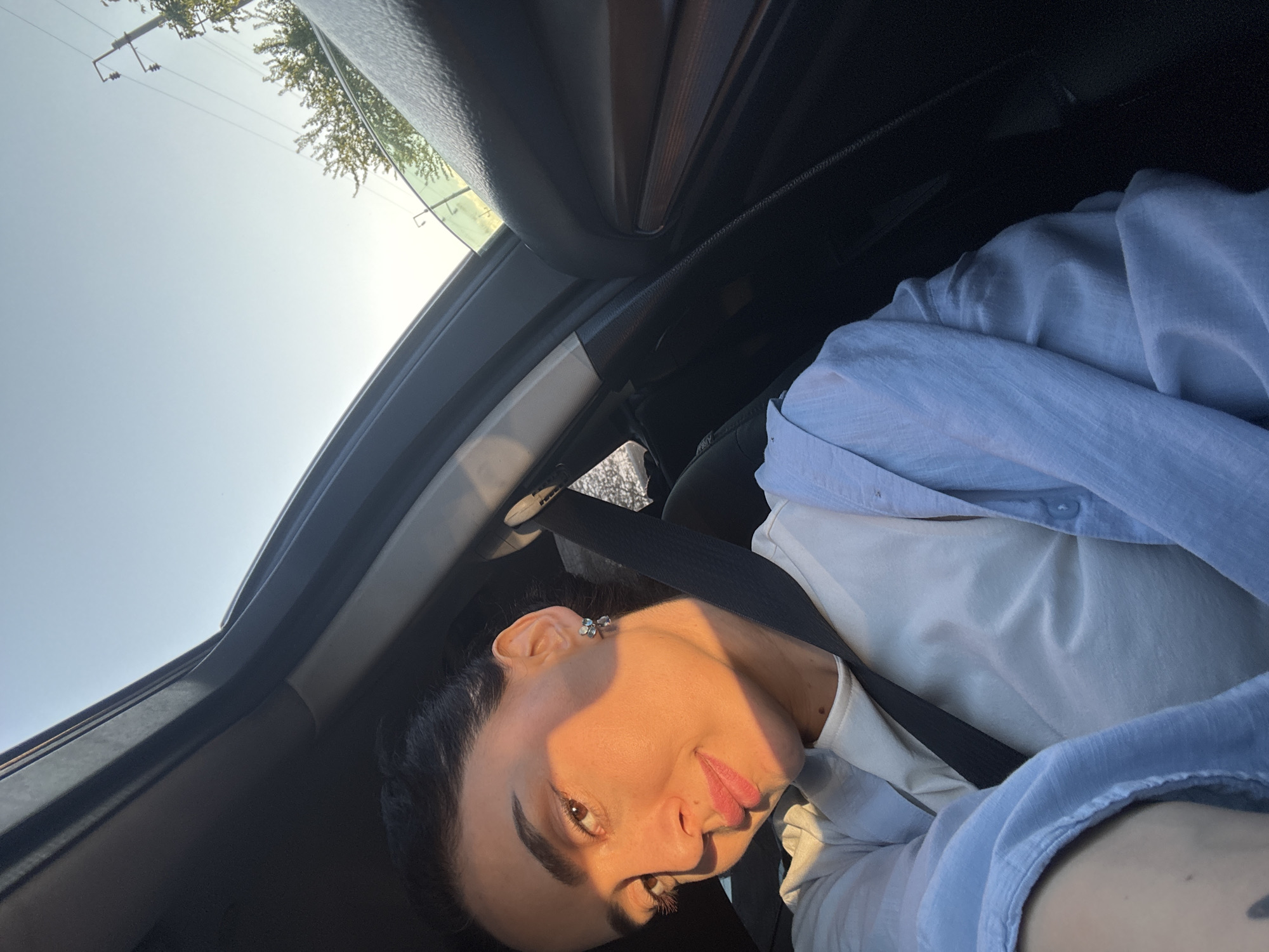 An overhead, tilted selfie captures a person inside a car, looking directly at the camera. The subject, likely a woman, has dark hair and eyebrows, brown eyes, and full lips, showing a subtle smile or smirk. Strong, warm sunlight illuminates the right side of her face, casting a distinct shadow across the left side, indicating a low sun angle, possibly late afternoon or early morning. She wears a white top underneath a light blue, long-sleeved collared shirt. A black seatbelt crosses her chest.
The car's interior is visible, including the dark headliner, window frame, and seat structure. Through the car's window or sunroof, a bright, clear sky is visible, along with glimpses of green tree foliage and power lines or utility poles against the blue. The scene suggests a sunny day. No text is visible in the image.