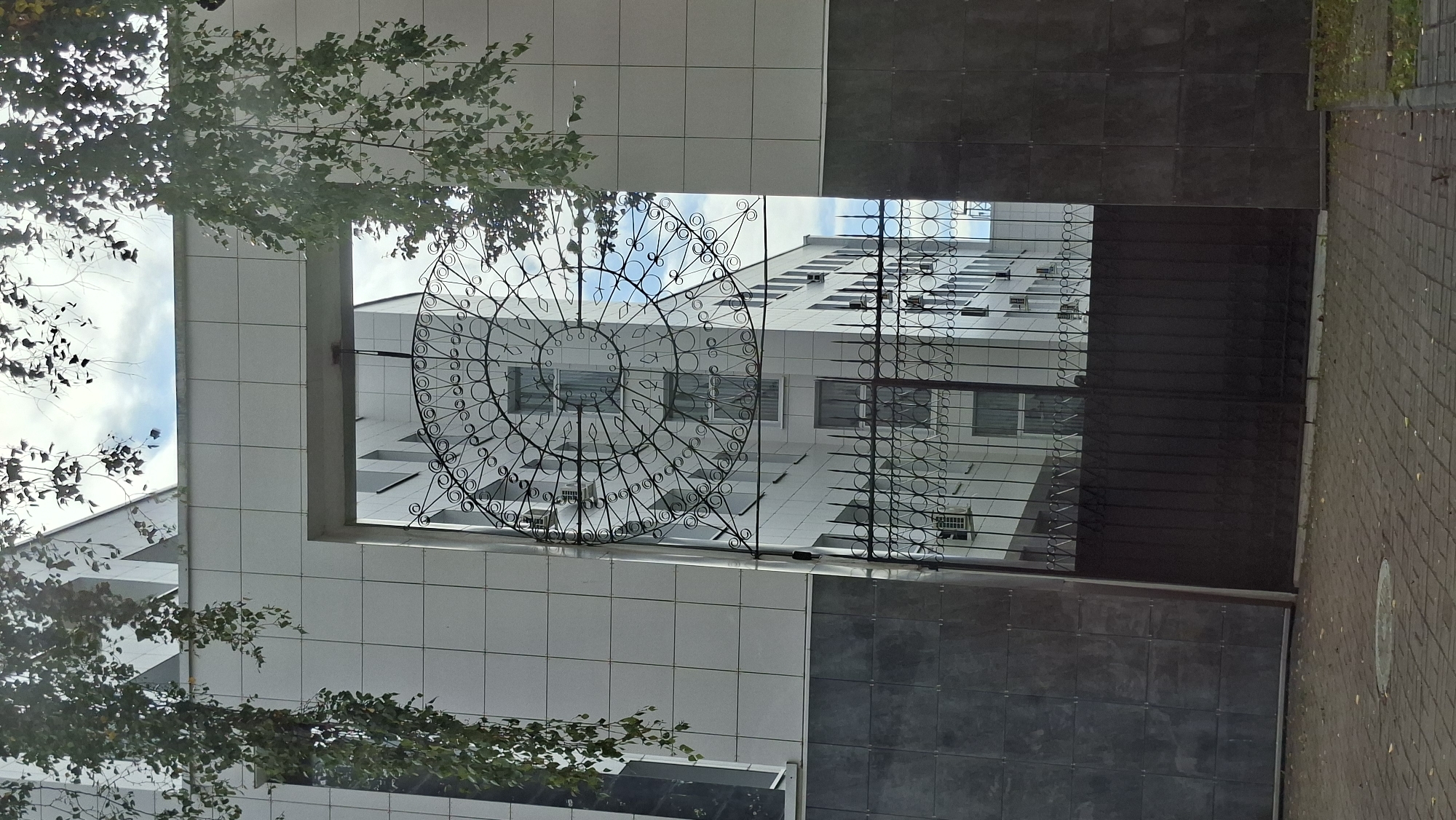 The image shows a view through an ornate wrought-iron gate towards a modern white tiled building. The gate is decorated with a circular pattern at the top, resembling a sunburst or a stylized flower. Behind the gate, the building features multiple windows and air conditioning units. Overhanging branches with green and brown leaves frame the top of the image, suggesting an outdoor setting, possibly in a city given the architecture. The foreground shows a paved area with cobblestones and some fallen leaves. The sky is visible through the gate and between the leaves, with some clouds. The overall impression is one of an entrance to a contemporary building, with a touch of decorative artistry provided by the gate. This scene is located in the city of Vitsebsk, Belarus, specifically at the entrance to the Vitebsk State Technological University.