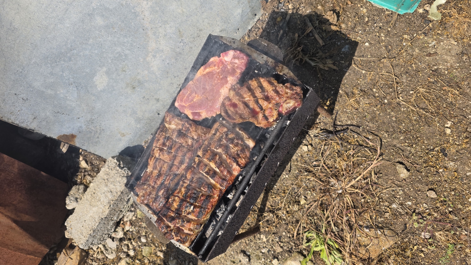 The image depicts a charcoal grill in an outdoor setting in Katuntsi, Bulgaria. The grill is rectangular and black, containing three pieces of meat: one raw, one cooked steak, and a rack of grilled ribs. Smoke is rising from the grill. The grill is situated on a ground with a mixture of dirt, dry grass, and small rocks. A concrete surface is visible to the left of the grill.