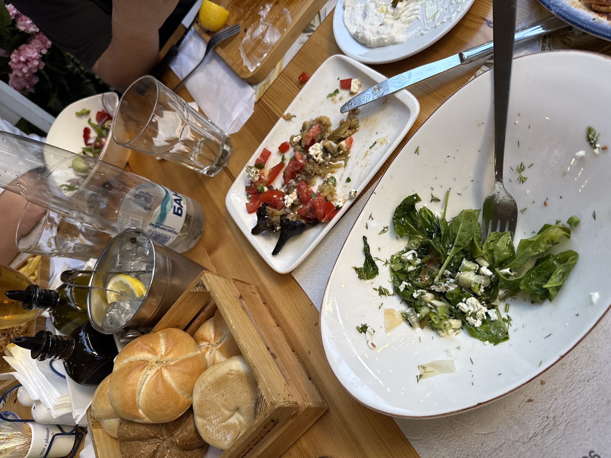 The image depicts a wooden table at a restaurant in Plovdiv, Bulgaria, likely outdoors given the presence of pink flowers in the background. The table is set with various partially eaten dishes and drinks, suggesting a meal has recently concluded or is in progress.
Dominating the foreground is a large white oval plate containing a partially consumed green salad with feta cheese and herbs, accompanied by a fork. To the right of this plate, a smaller white oval plate holds a white dip or spread, also partially eaten. Further to the right, a rectangular wooden board shows remnants of food and a lemon wedge.
In the middle ground, a white rectangular plate holds a partially eaten dish of roasted eggplant with tomatoes and feta cheese, alongside a knife. Behind this, a wooden crate contains several rolls, some whole and some halved. Beside the crate, a silver bucket is filled with ice and a lemon slice. Several glasses are visible, including an empty clear glass and a plastic water bottle with a blue label.
In the background, two dark glass bottles, likely containing olive oil and vinegar, are positioned behind the ice bucket. A person's arms and torso are visible on the right side of the image, dressed in a dark shirt, indicating their presence at the table. The overall scene is casual and indicative of a dining experience.