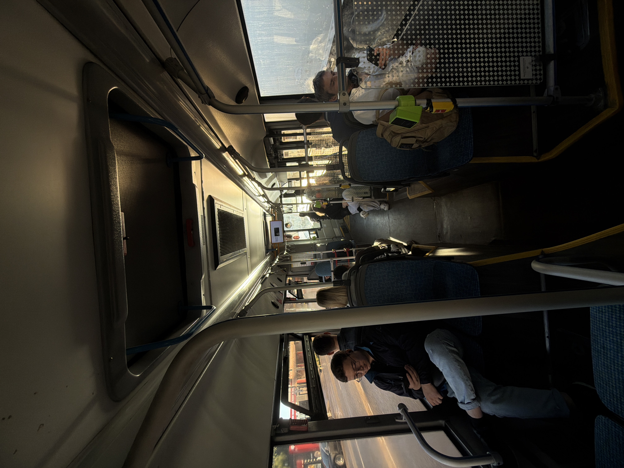 An interior view from a bus in Butovo, Russia, shows passengers seated as the bus travels. The light filtering through the windows indicates it is daytime, with the sun casting long shadows. In the foreground, a man wearing glasses and a dark jacket sits on the left, holding a smartphone. Across the aisle, another man in a light-colored shirt is also looking at his phone. Several other passengers are visible further down the bus, some facing forward and others looking towards the windows. The bus interior features blue patterned seats, metal support poles, and overhead handrails. The windows offer a view of a street with buildings and a car passing by.