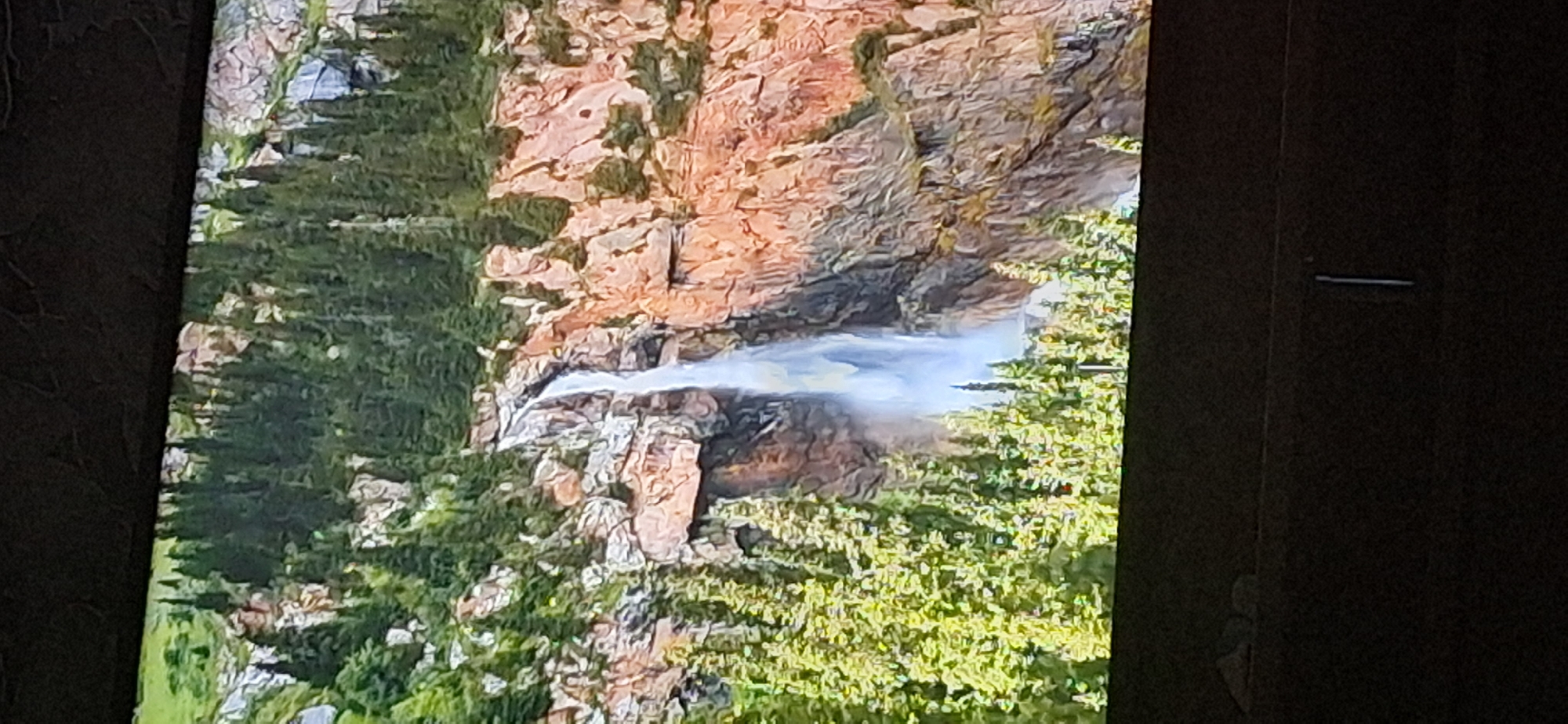 The image shows a waterfall cascading down a rocky, mountainous terrain. Lush green pine trees are visible on the slopes, both above and below the waterfall. The rocks appear to be a reddish-brown color, with some lighter gray and white sections. The sky is not visible in this frame. The scene is depicted on what appears to be a screen, as evidenced by a dark border at the top and bottom of the image, and a textured surface above the screen. The location of this scene within the city of Bila Tserkva, Ukraine, is not discernible from the image itself.