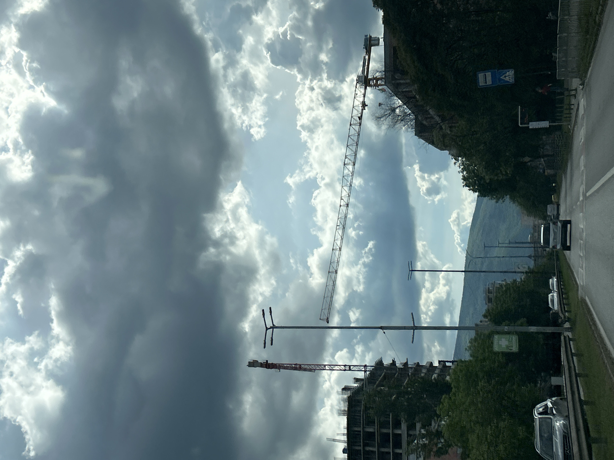 The image shows a street scene with a partially constructed building and a crane visible against a cloudy sky. A road with vehicles is on the right side of the frame, lined with trees and street lights. A mountain is faintly visible in the background. The exact location in Sofia, Bulgaria, cannot be definitively determined without additional information or landmarks.