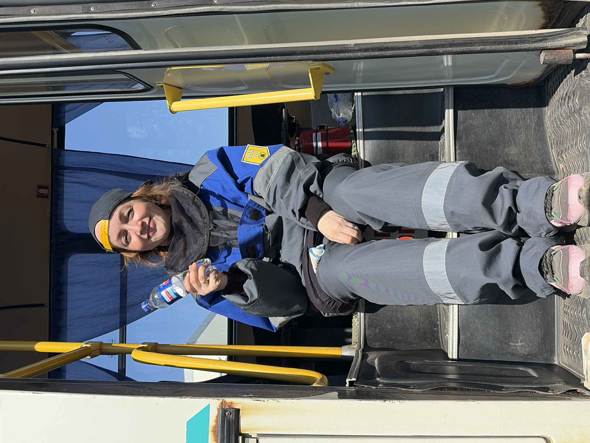 A woman wearing a blue and grey work uniform with reflective stripes is sitting on the steps of a bus. She is holding a bottle of water in her hands. The location is Opary, Russia. The bus has yellow handrails. There is a fire extinguisher mounted on the wall behind her.