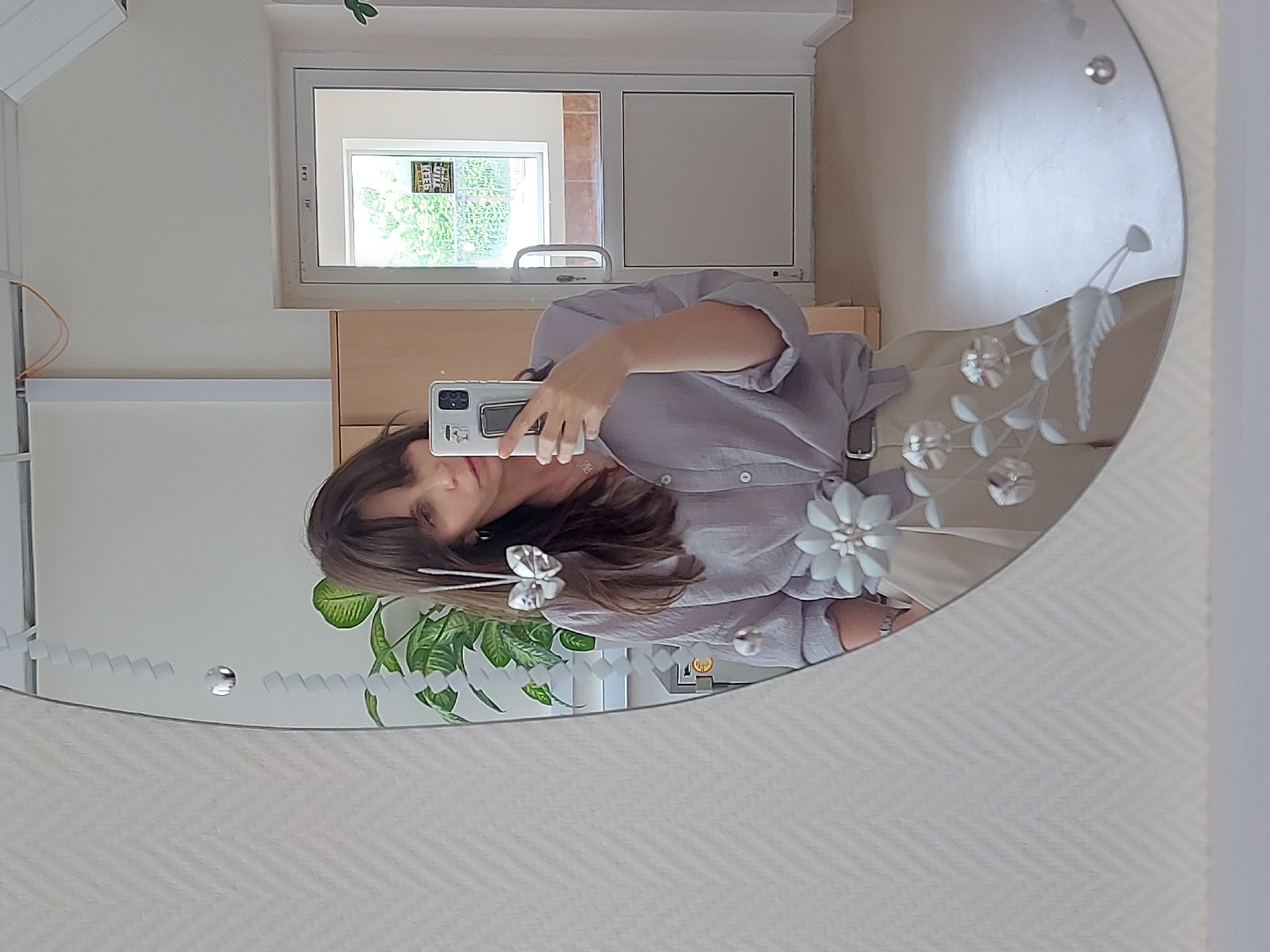 The image is a mirror reflection of a woman taking a selfie in what appears to be an indoor setting. The woman is wearing a light grey button-down shirt, and her brunette hair is adorned with a white bow clip. She holds a smartphone with a visible camera lens in her left hand, capturing her own reflection.
The mirror's design features etched floral patterns along the edges. Reflected in the background is a partially visible interior: a closed white door with a window providing a view of greenery and possibly an adjacent building, wooden cabinets, and a potted plant.
Based on the reflection, the wall behind the mirror has a neutral tone with a faint textured pattern. No specific details indicate the location as Chaikovsky, Russia, or any other particular city.
