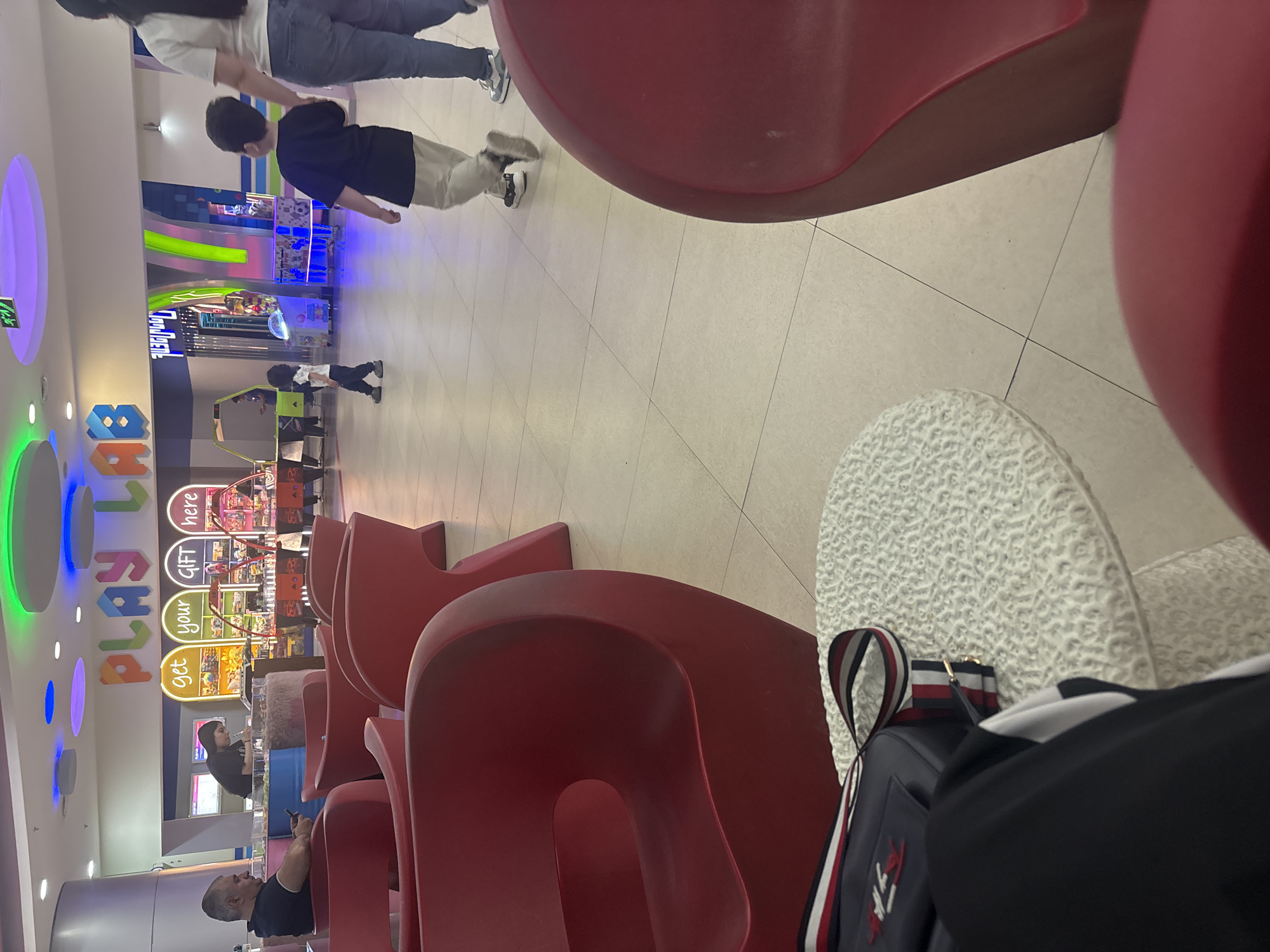 The image depicts an indoor scene within a modern entertainment or shopping mall in Yerevan, Armenia, specifically focusing on a play area called "PLAY LAB." The photograph is taken from a low perspective, looking across a row of red, contemporary seats. In the immediate foreground on the right, part of a white, textured chair or stool is visible, alongside a black bag adorned with red, white, and blue stripes, suggesting a casual setting.
In the mid-ground, a man in a black polo shirt is seated on one of the red chairs, engrossed in his phone. Behind a counter to his left, a woman with dark hair stands, appearing to work or attend the area. The background showcases a vibrant arcade or amusement zone with several arcade machines and bright neon lighting in shades of blue, green, and purple. Several individuals are present in this area: a woman in a white top and jeans walks away from the camera with a child in a black shirt and light pants, while another small child in black is seen near an arcade machine.
Prominently displayed on the left wall of the play area are colorful, illuminated letters spelling out "PLAY LAB." Above a counter in the mid-ground, signs in yellow and orange read "get your GIFT here." The floor is a light-colored tiled surface. The scene suggests a lively, leisurely atmosphere typical of a family-friendly entertainment venue, likely during the daytime or early evening given the bright, consistent indoor lighting.