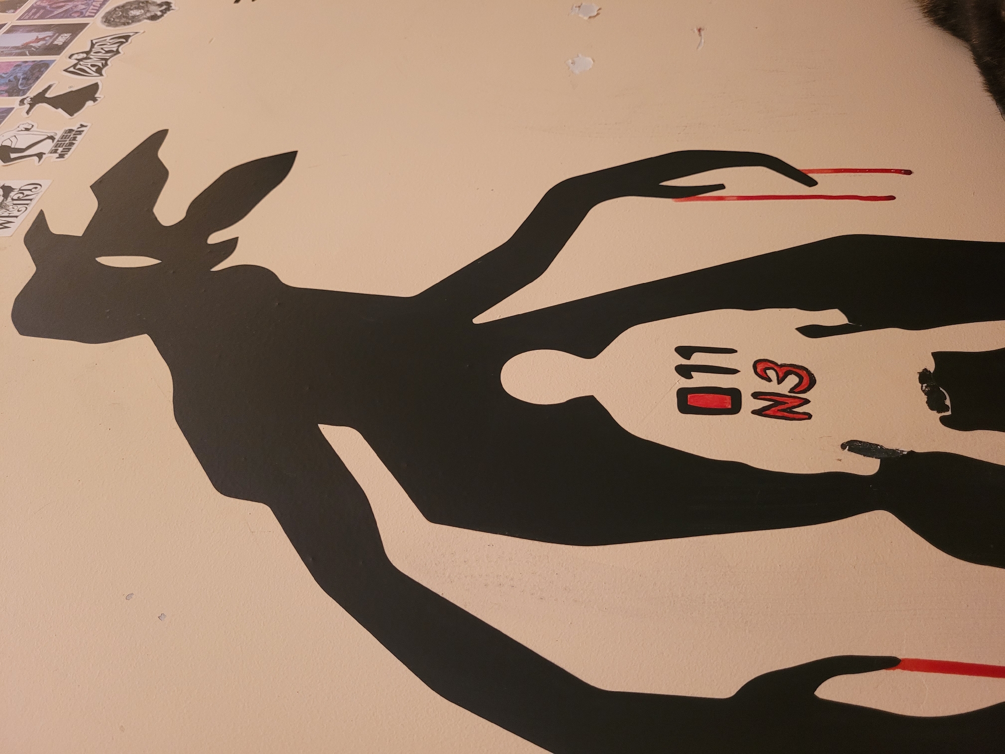 On a light tan or beige wall in what is potentially Izhevsk, Russia (given the Cyrillic "ИЗ" visible), there's a black silhouette of a creature reminiscent of a deer or jackalope with antlers, but the form is more abstract. In the negative space within the silhouette's body is an outline of a small human figure, and near this figure, the stylized letters "ИЗ" are painted in red and black. Extending from the silhouette are two arms, and at the end of each arm, a hand. Between the fingers of the hands are two parallel, horizontal red lines, appearing like chopsticks or perhaps paintbrushes. At the top left corner of the image, a series of stickers are visible, suggesting a casual or decorated environment. The wall seems to have some minor damage, such as peeling paint.