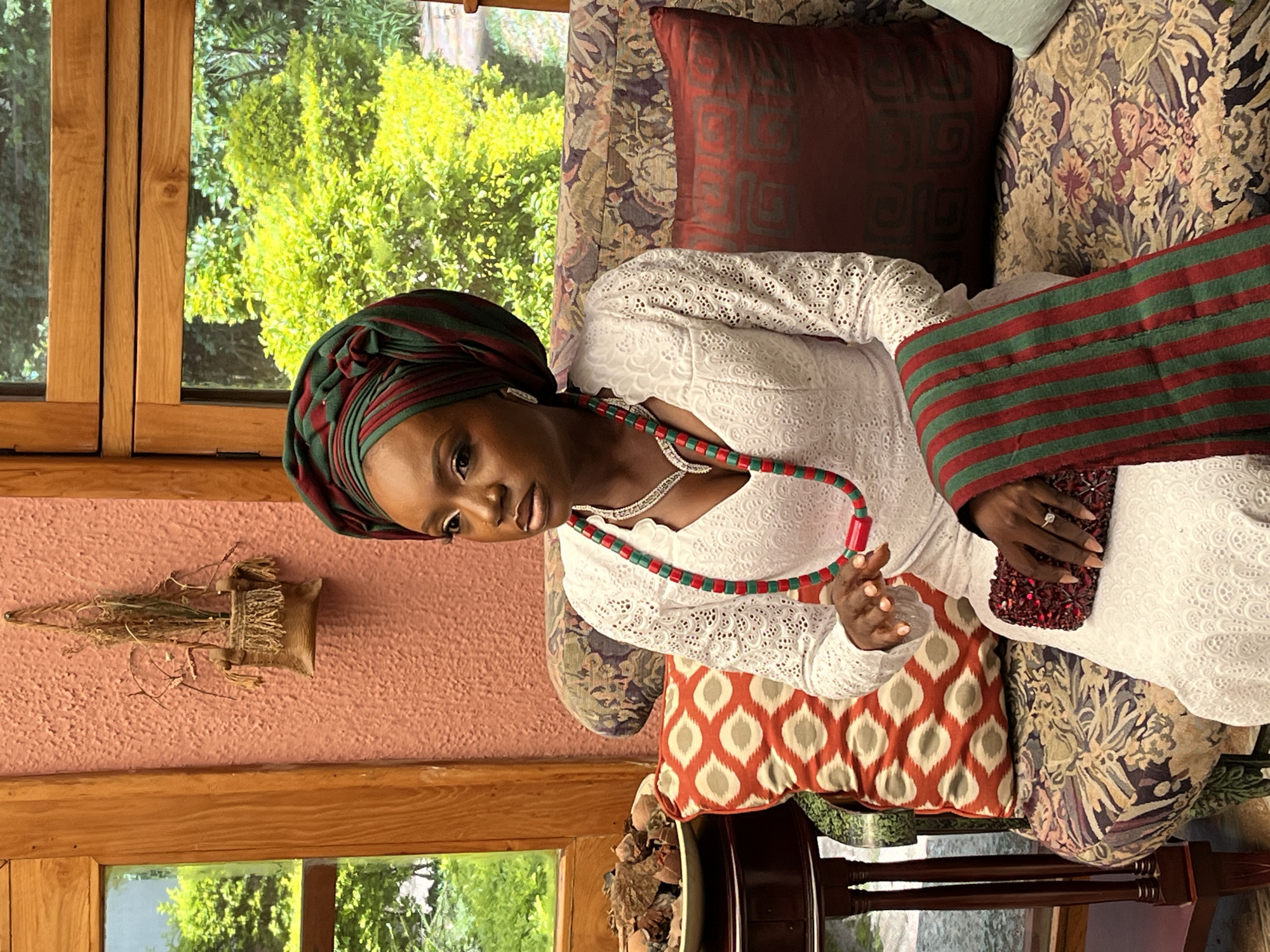 A Black woman, adorned in a white lace dress, a vibrant green and burgundy headwrap, and a matching beaded necklace, is seated on a patterned sofa in what appears to be a home in Bar Jirgi Summa, Nigeria. She holds a small, jeweled clutch bag in her lap and a striped scarf is draped over her arm. The room features a textured pink wall, large wooden-framed windows revealing lush green foliage outside, and decorative pillows on the sofa. A small, woven basket with dried plants hangs on the wall to the left of the woman. A round side table to her left holds a bowl filled with dry, natural materials. The overall setting suggests a comfortable, possibly traditional, interior space.