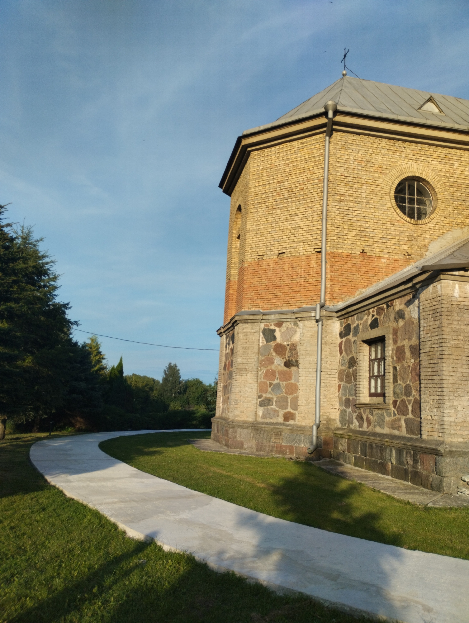 A concrete pathway curves through a grassy area beside a stone and brick building under a clear blue sky. The building, which appears to be a church, has a light yellow brick facade with a band of red brick near the top. A circular window with a cross-shaped lattice is visible on the upper section of the building. The lower portion of the building is constructed with irregular, multi-colored stones. A small, dark-framed window is present on this stone section. A metal downspout runs vertically along the side of the building. Tall evergreen trees are located to the left of the building, and a line of deciduous trees forms a backdrop. The sun casts shadows from the trees onto the pathway and grass. No people are visible in the image. The scene is located in the vicinity of Вішнеўскі сельскі Савет, Belarus.