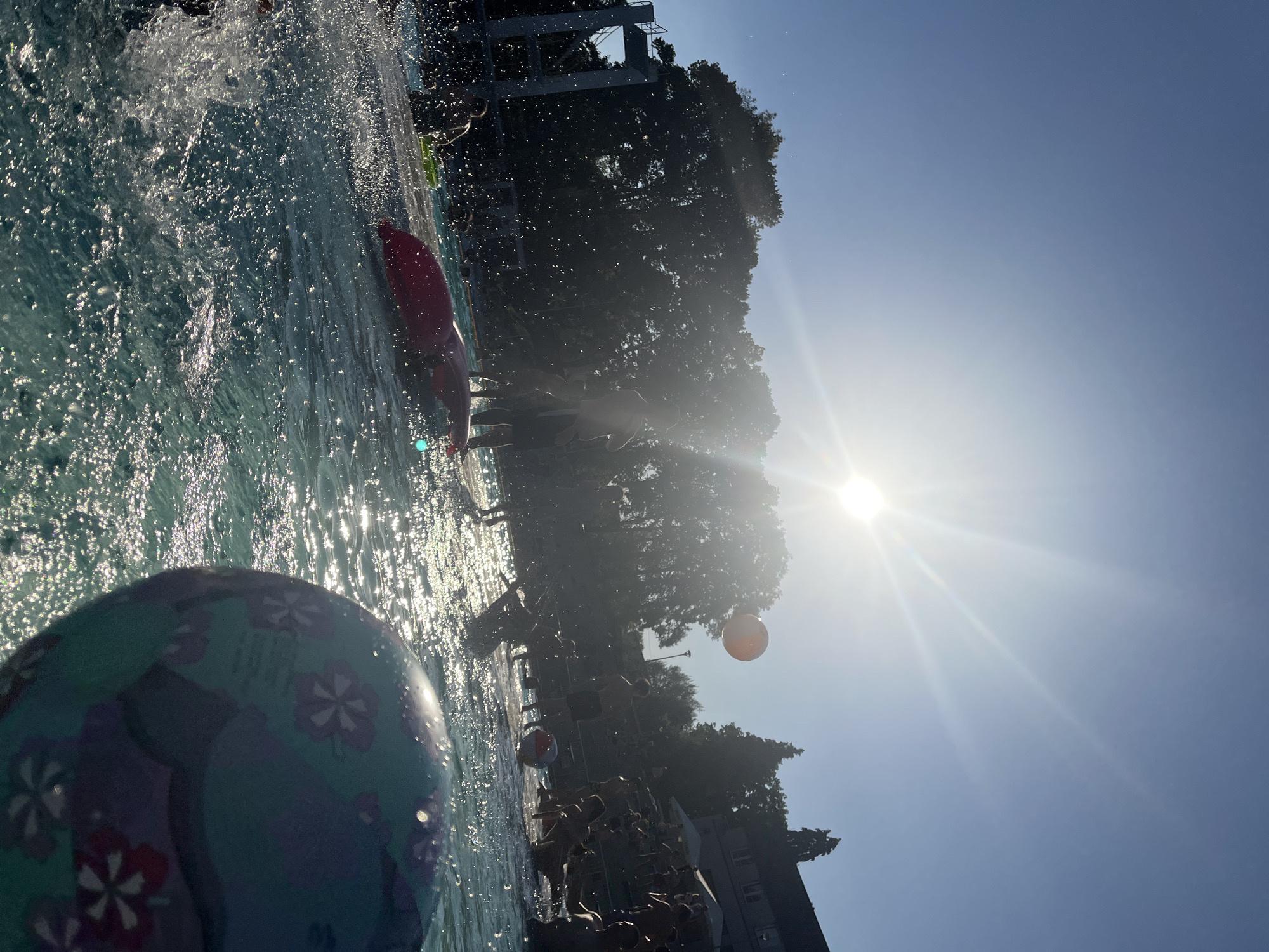 This is a tilted, close-up, underwater shot of a swimming pool on a sunny day. The water is clear and blue, with sunlight glinting off the surface, creating a shimmering effect. The image is dominated by the water and the sky, with a bright sun shining intensely in the upper right quadrant. Silhouettes of trees and buildings are visible in the background, along with a diving platform.
Several people are in the pool, mostly appearing as dark shapes due to the lighting conditions. One person is visible wearing a red inflatable ring. There are also what appear to be inflatable balls floating in the water. In the foreground, a large, round object with a floral pattern, possibly a beach ball or a hat, takes up a significant portion of the frame. The overall impression is one of a fun, recreational day at a pool, with people enjoying themselves in the water under a clear sky. The sun suggests it's daytime, likely midday or afternoon.