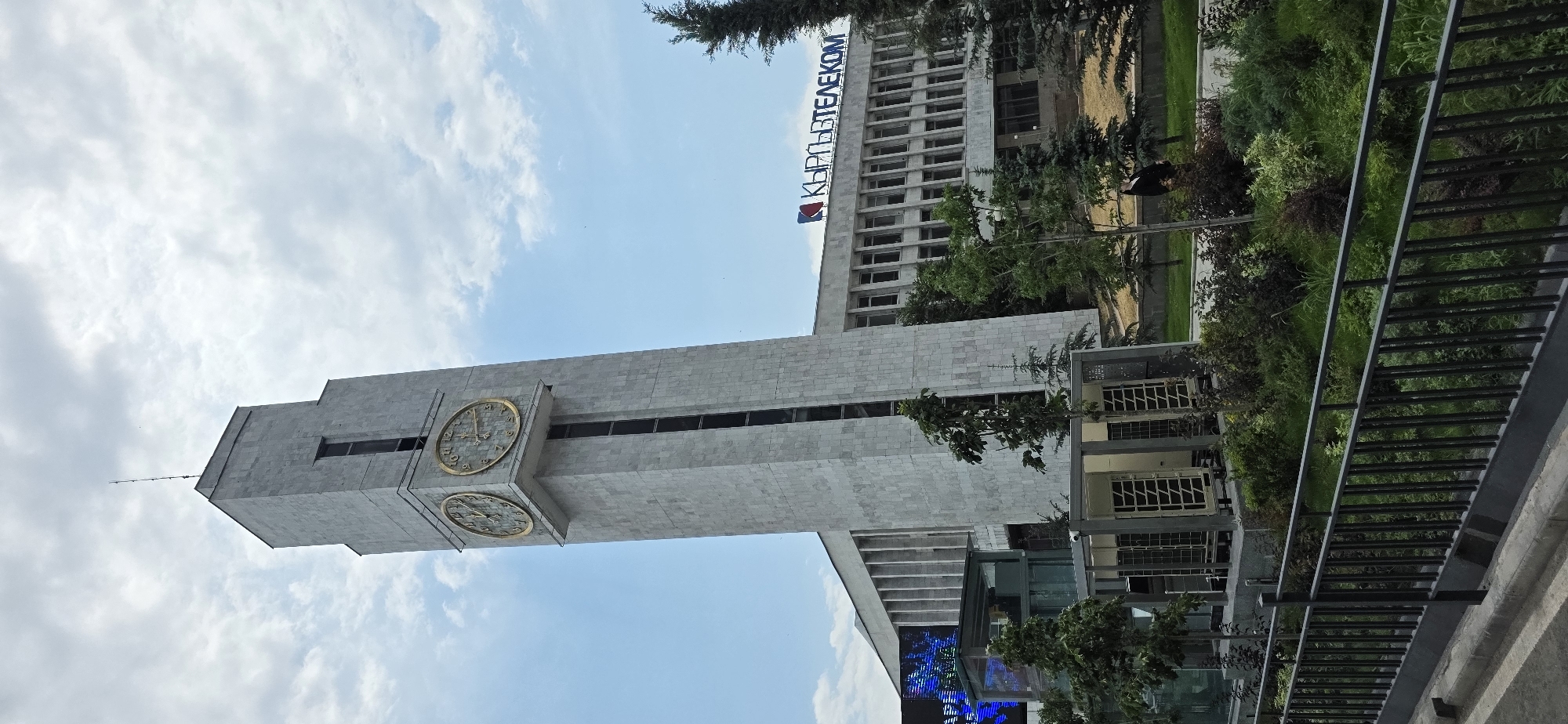 A tall, concrete clock tower with two large, gold-faced clocks is prominently featured in the center of the image, leaning slightly to the left. The tower is clad in light-colored stone tiles. Below the clock tower, a modern, rectangular building with many windows stretches horizontally across the frame. A blue and red logo with Cyrillic lettering, reading "КЫРГЫЗТЕЛЕКОМ" (Kyrgyztelecom), is visible on the top facade of this building. In front of the building and around the base of the clock tower, there is a landscaped area with various trees and bushes. A dark metal fence runs across the foreground of the image, separating it from the sidewalk and a sloped curb. The sky above is partly cloudy, with patches of blue visible. To the left of the clock tower, a large LED screen displaying a blue, abstract pattern is partially visible. A person in a dark jacket is standing in the middle ground, to the right of the building. The image appears to be taken from a street-level perspective.