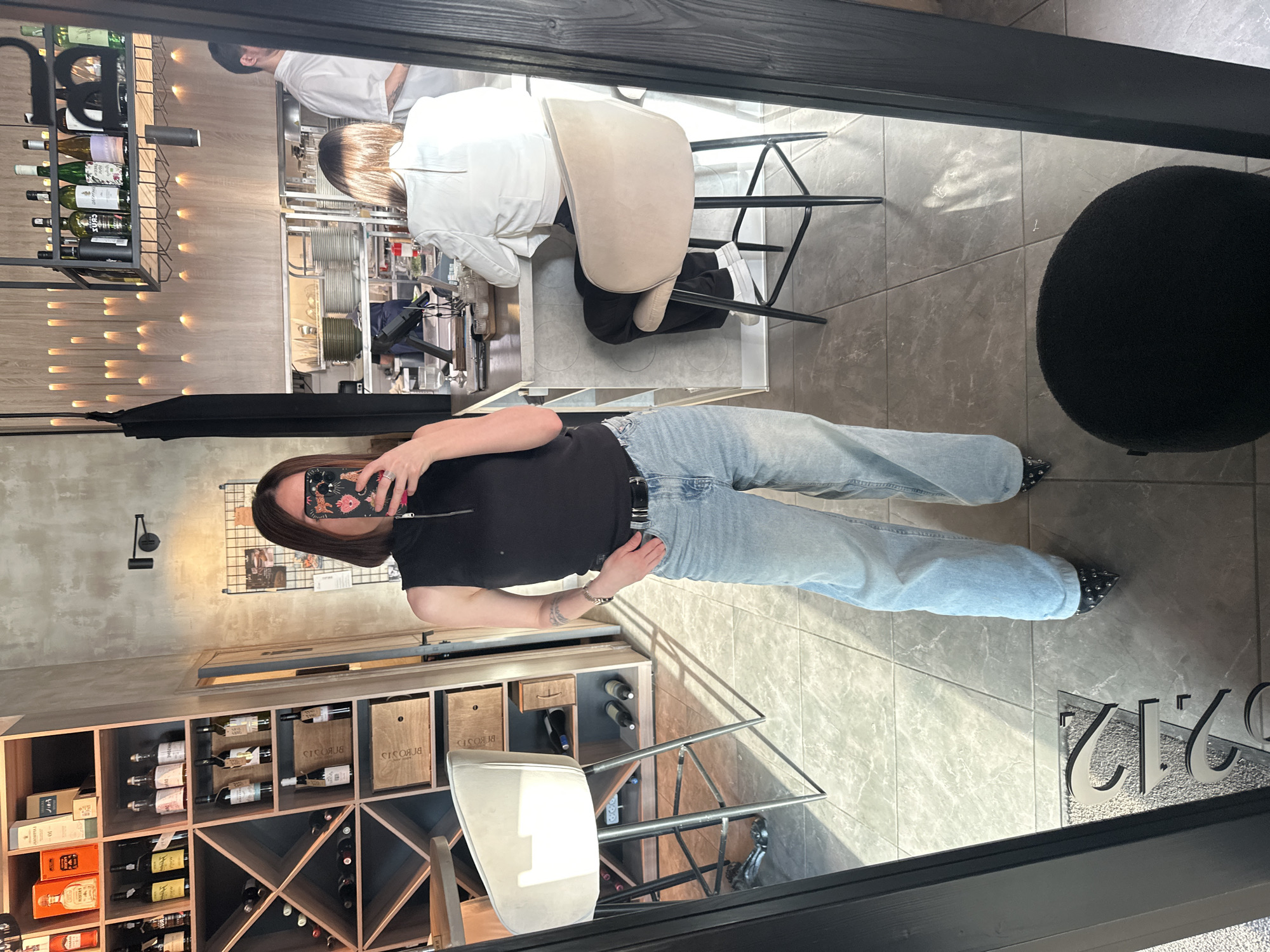 A selfie taken in the 212 Bar in Izhevsk, Russia, shows a woman in a black top and light blue jeans standing in front of a wine rack. The wine rack is filled with wine bottles and wooden boxes. The woman is holding her phone up to take the photo. In the background, chefs are visible behind a counter, preparing food. A black, round stool is partially visible in the reflection of the window. The floor is tiled in gray.