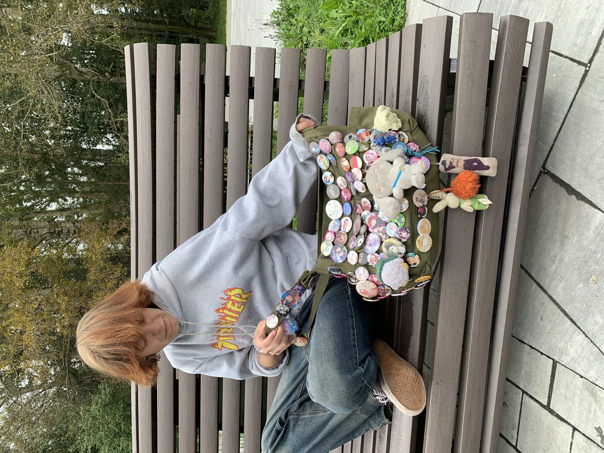 A person with short, reddish-blonde hair sits on a wooden park bench, looking down at a green messenger bag covered in numerous pin-back buttons and a few small plush toys. The person wears a grey hooded sweatshirt with a flame logo and blue denim jeans. One shoe, a tan and white sneaker, is visible. The bench is made of dark brown wooden planks and is situated outdoors on a paved area with some greenery and trees in the background. The lighting suggests it is daytime. The location of Prokopyevsk, Russia, is not ascertainable from the visual information in the image.