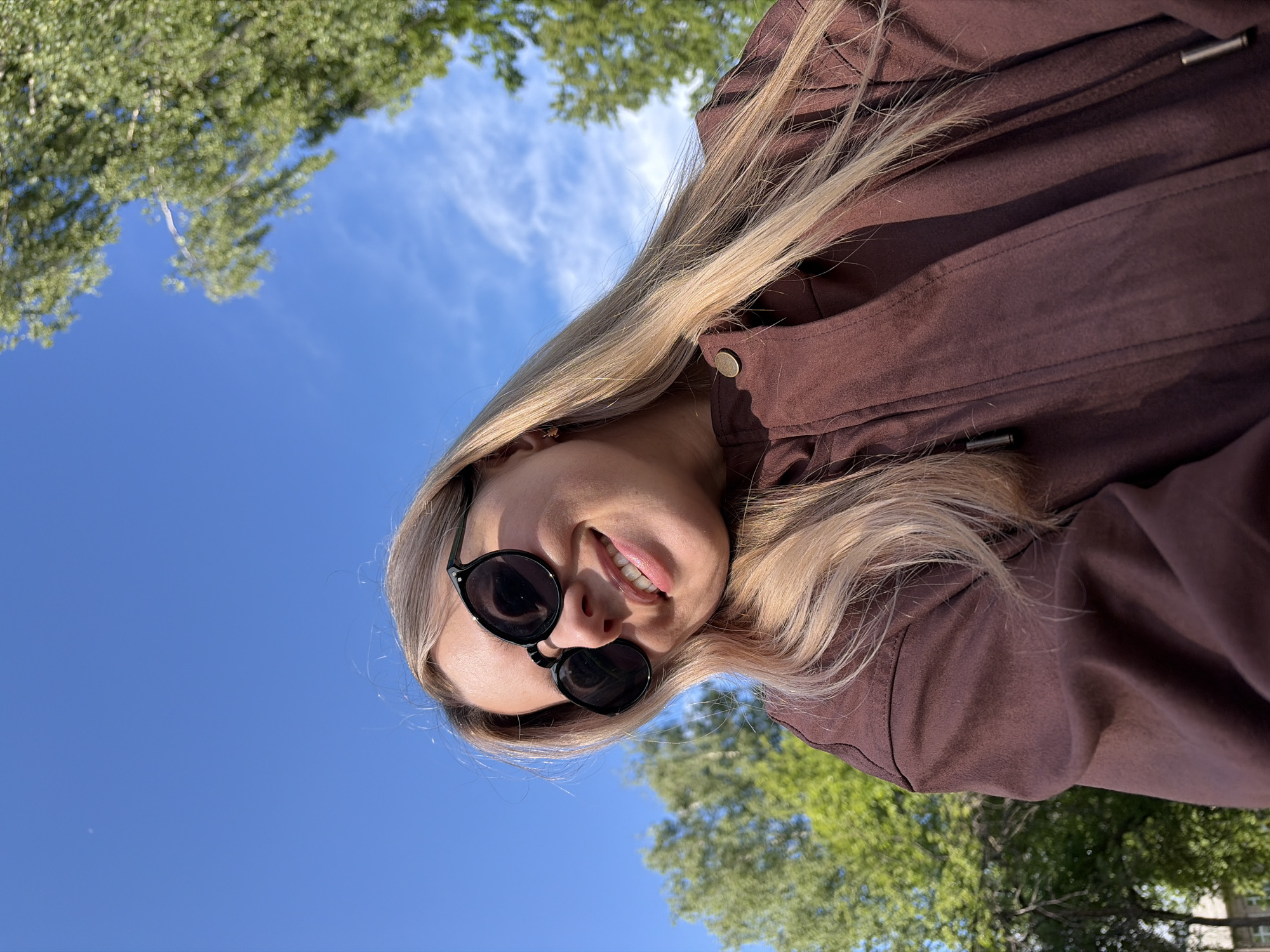 A young woman with blonde hair is posing for a selfie outdoors in Chaikovsky, Russia. She is wearing sunglasses and a brown jacket. The background features a blue sky with some clouds and green trees. The photograph is taken from a low angle, looking up at the woman.