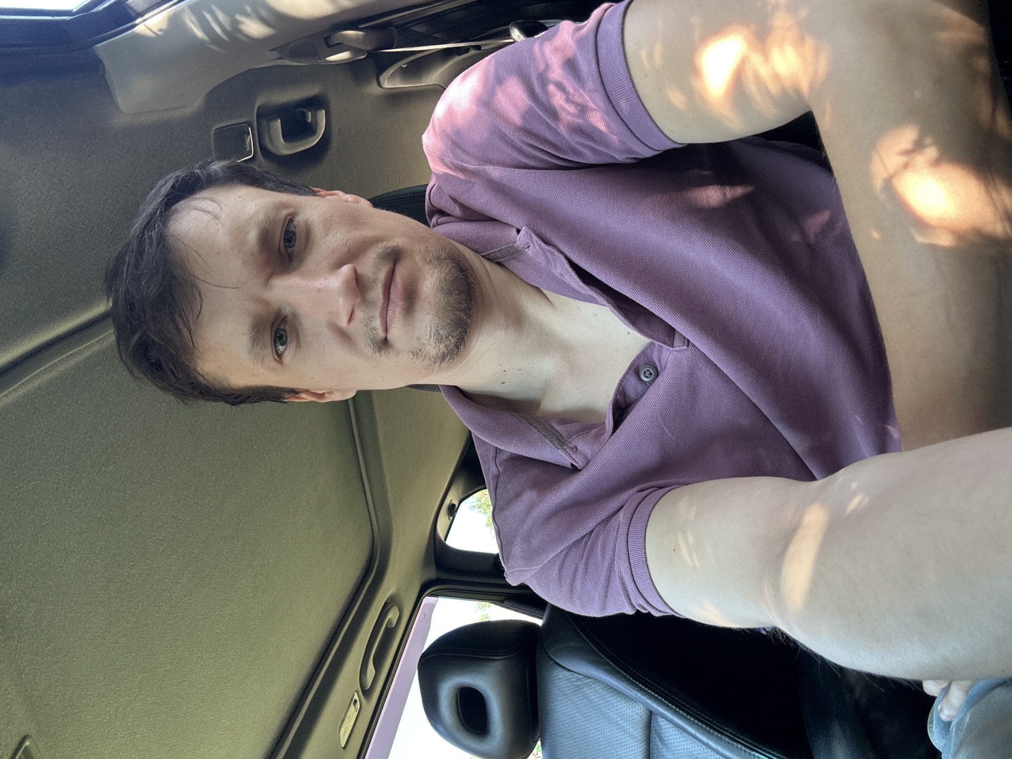 A man is seated in the driver's seat of a car, with the camera positioned for a selfie. The car's sunroof is open, and natural light streams in, casting dappled shadows on the man and his clothing. He is wearing a purple polo shirt. The car interior is visible, including a black leather seat and part of the dashboard. The scene appears to be outdoors, with glimpses of greenery visible through the open sunroof and car window. The location is specified as Shatava, Ukraine.