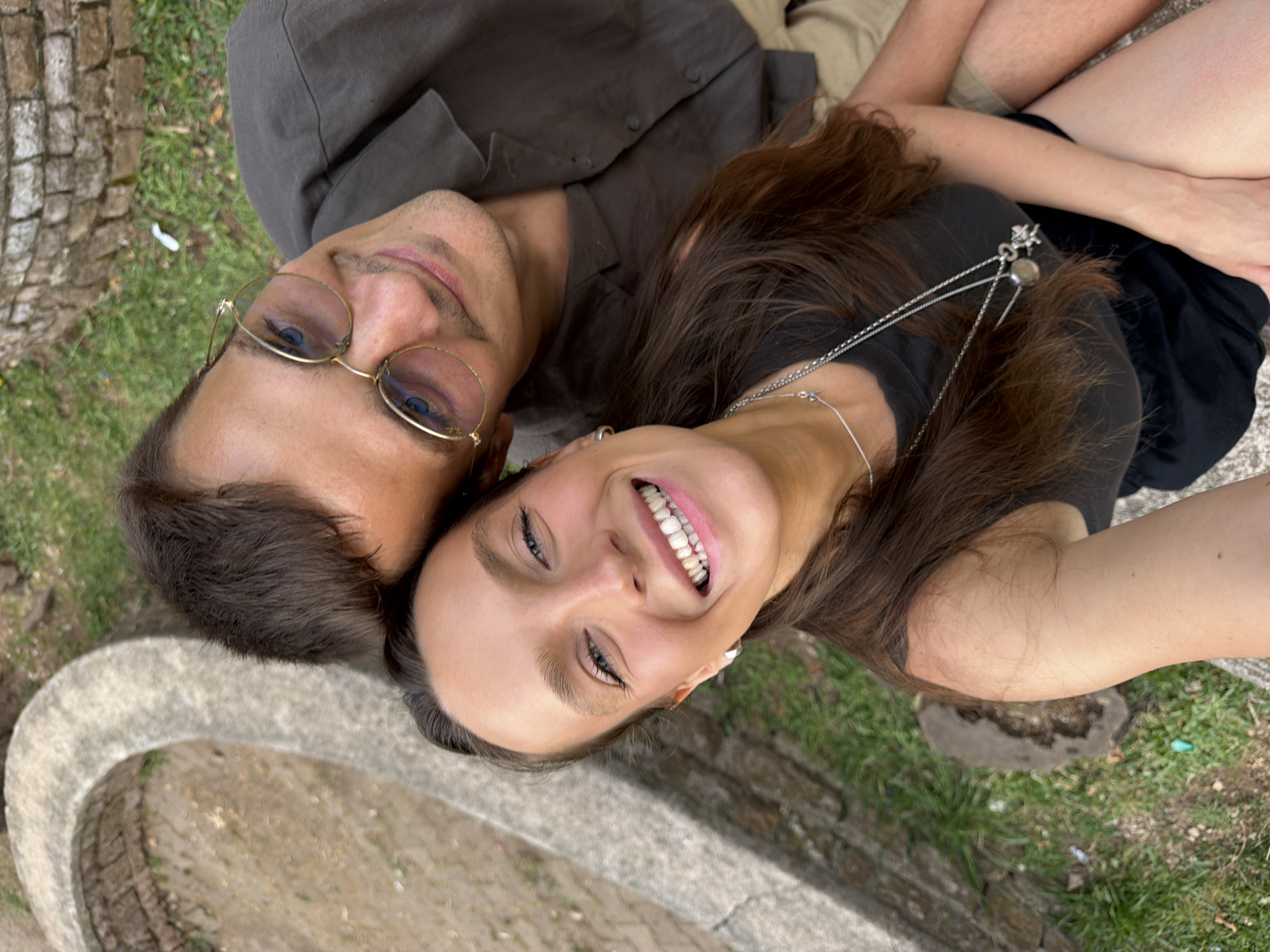 An eye-level, high-angle, close-up selfie shows a Caucasian man and a Caucasian woman from the chest up, posing outdoors in what appears to be a park or garden area. The man, on the right, is wearing a dark grey collared shirt and tan shorts. He has short brown hair and is wearing round, gold-rimmed glasses. The woman, on the left, has long brown hair and is wearing a black tank top and black shorts. She is wearing two silver necklaces, one with a circular pendant and the other with a small charm. Both individuals are smiling at the camera. In the background, to the left, is a curved stone structure, possibly part of a fountain or seating area, with some cobblestone visible underneath. The ground is covered in green grass and some dirt. The overall lighting suggests it is daytime. The presence of the curved stone structure, the cobblestone pathway, and the grassy areas are consistent with public spaces often found in historic city centers or parks. Given the context of Budva, Montenegro, this scene could be located within the Old Town's vicinity or one of its public gardens.