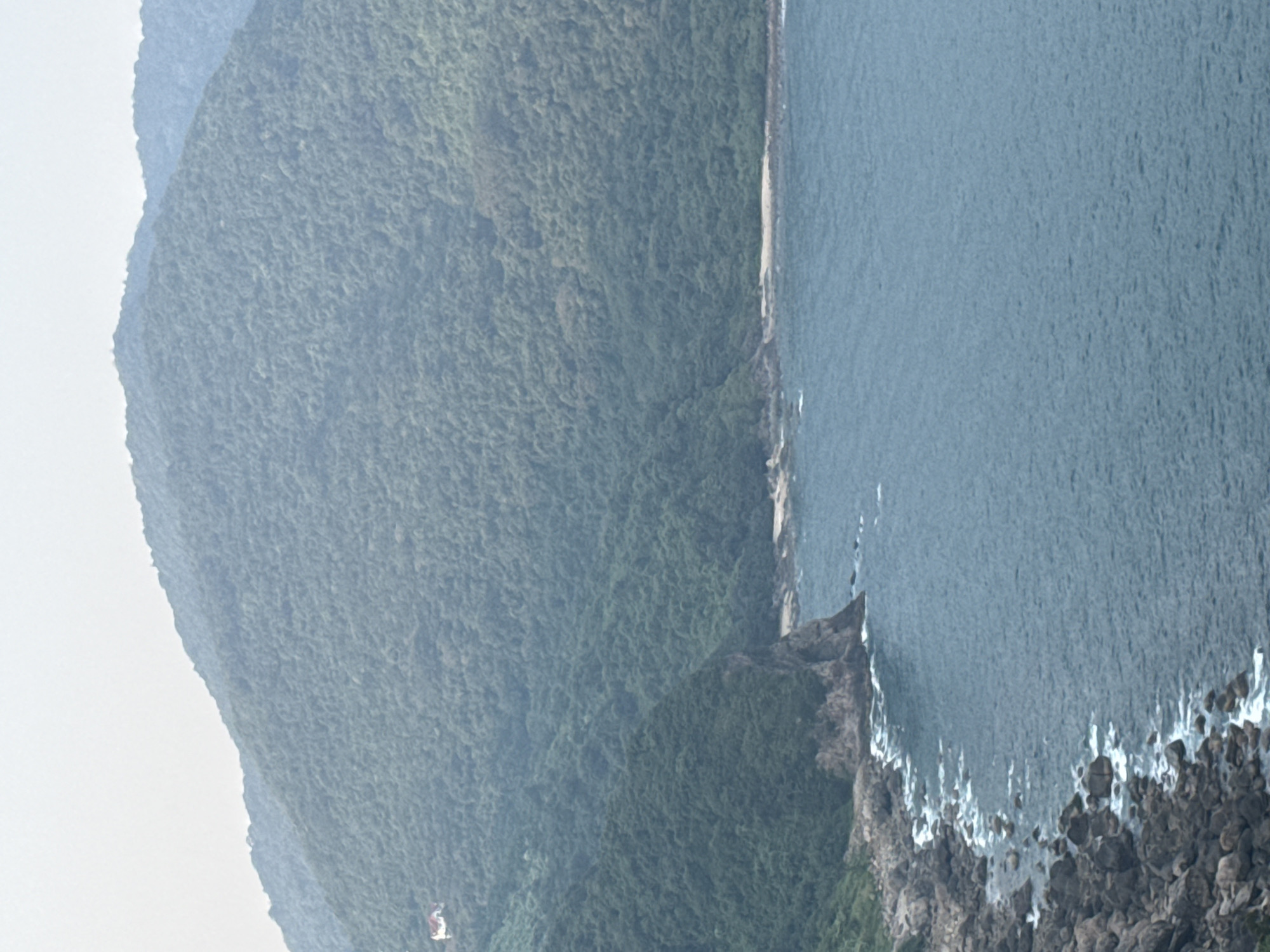 The image depicts a mountainous coastline with lush green vegetation along the slopes. The sea, characterized by a moderate hue of blue, occupies the right side of the view with white waves crashing against a rocky shore in the foreground. The terrain is steep and rugged.
While the image lacks identifiable landmarks or specific details to pinpoint the precise location, it aligns with the coastal landscape often observed in the Sơn Trà Peninsula region of Đà Nẵng, Vietnam, where mountains meet the sea. There are no discernible people, subjects, objects, actions, or interactions present within the frame. The environment is predominantly natural, dominated by the sea and vegetated mountain range.