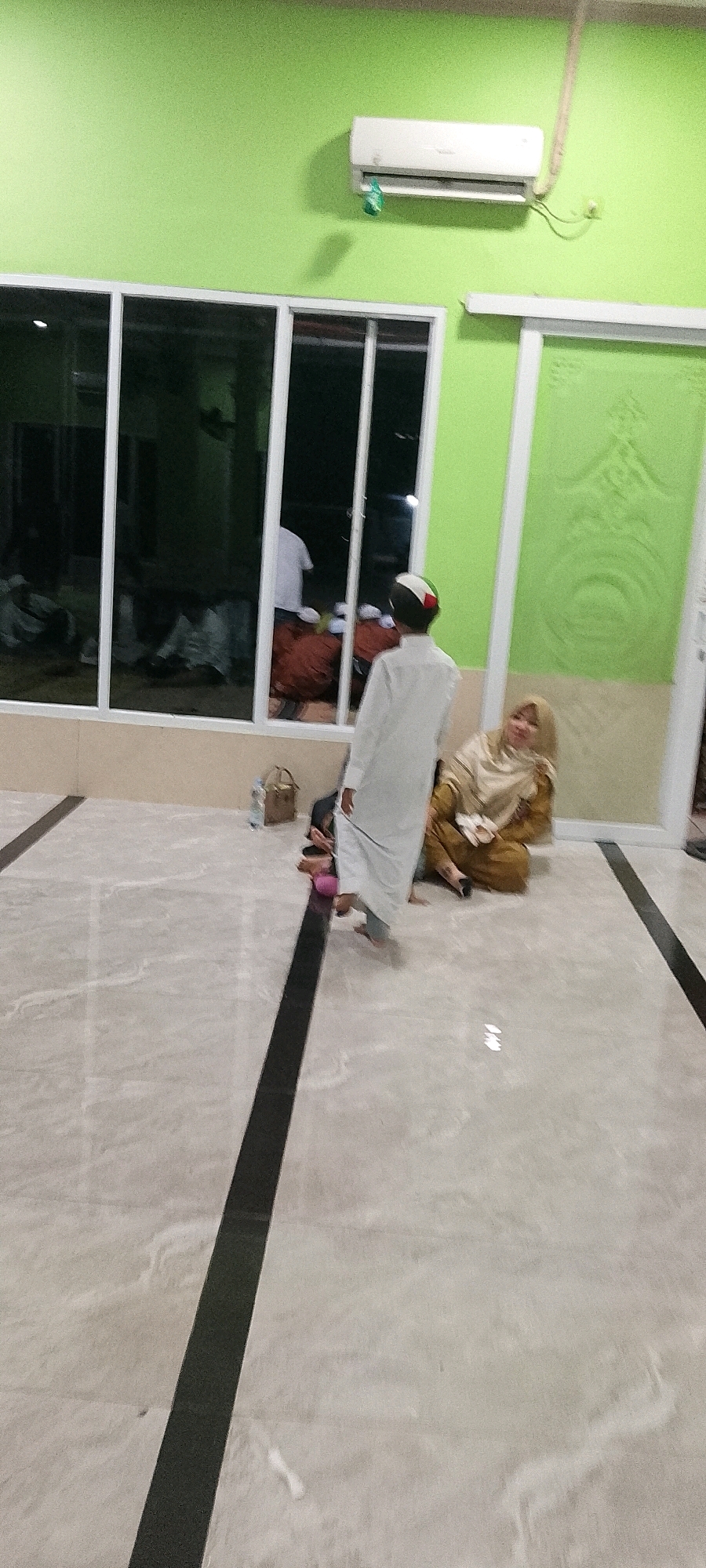 The image depicts an indoor scene, likely a prayer room or gathering space, with a light green painted wall and large windows. Inside the windows, several individuals are seen in prayer or sitting down. Outside the windows, in the foreground, a person wearing a white thobe and a patterned kufi walks away from the camera. To the right, a woman in a beige hijab and matching dress is seated on the floor, holding a pair of sandals. A brown woven bag and a bottle of water are on the floor near her. The floor is tiled with a light marble pattern and features two black linear inlays. An air conditioning unit is mounted on the upper part of the green wall. The image is taken from a low angle, looking across the floor and up towards the wall and windows. The overall lighting suggests an indoor environment, possibly at dusk or with artificial lighting. The location is identified as Satriamekar, Indonesia.
