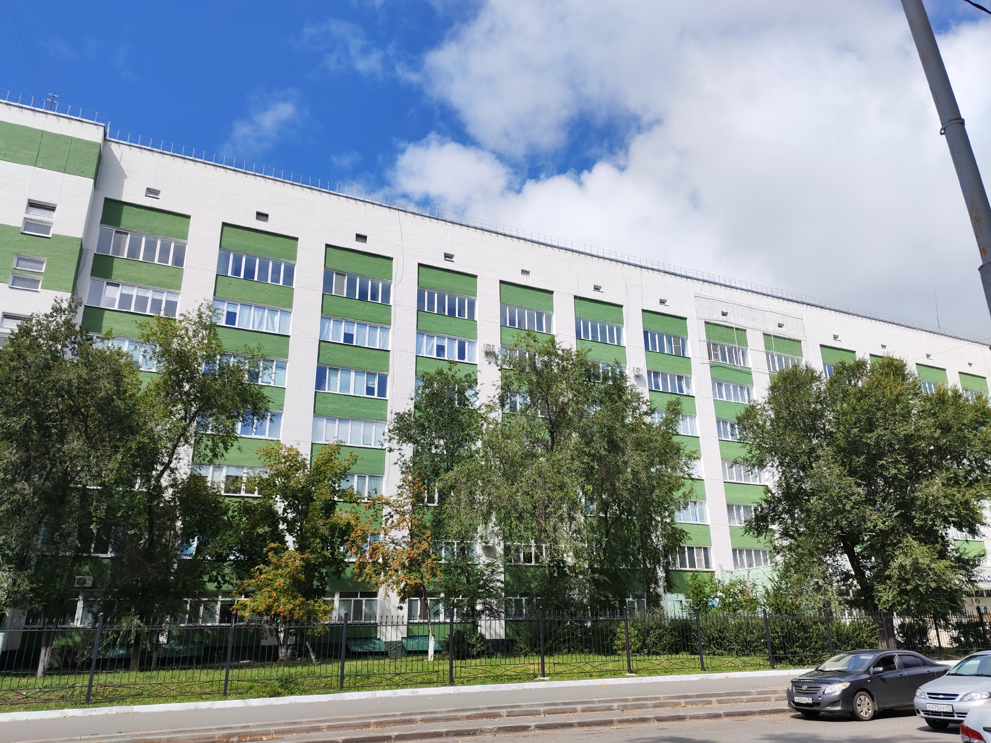A long, multi-story building with white and green facade is seen from a low angle. The building has many windows with white frames. Trees with green foliage are in front of the building. A black fence separates the trees from a sidewalk. Two cars are parked on the sidewalk. The sky is blue with some white clouds.
