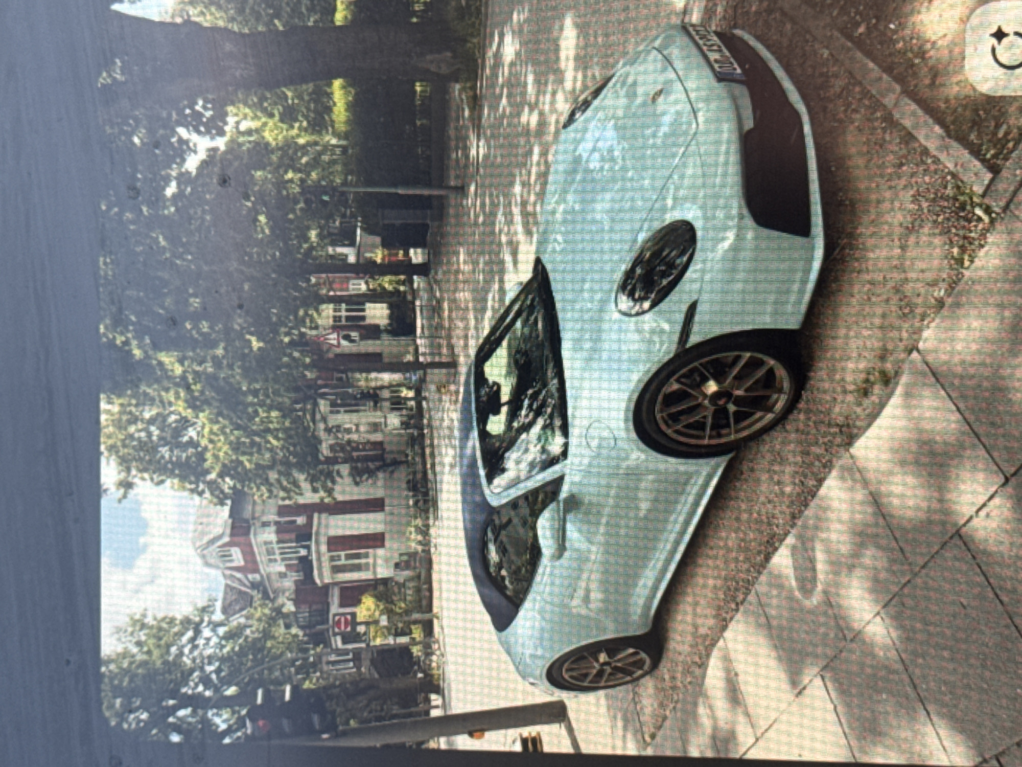 The media file displays a light blue or mint green sports car, resembling a Porsche 911, parked on a paved surface. The image is oriented vertically, showing the car positioned diagonally across the frame, with its front facing towards the top-left and its rear towards the bottom-right. The car features dark, multi-spoke wheels. Adjacent to the car, on its right side, is a patterned sidewalk made of light-colored tiles.
The scene is an outdoor urban or suburban environment, bathed in bright daytime sunlight, which casts distinct shadows and highlights on the ground. A dense canopy of green trees fills the left side and background of the image, contributing to the dappled light effect. Beyond the trees, several multi-story buildings are visible, characterized by red-tiled roofs and white facades with red accents, suggesting a residential area. A small, circular red and white traffic sign is partially visible in the distance.
No people are present in the image, and the car appears stationary. The rear license plate of the vehicle clearly displays the text "C 8857 TO". This alphanumeric sequence is consistent with a Bulgarian license plate from the Varna province, confirming the location context of Varna, Bulgaria. In the top-right corner of the image, a white user interface icon, possibly a smiling face or camera symbol, is visible.