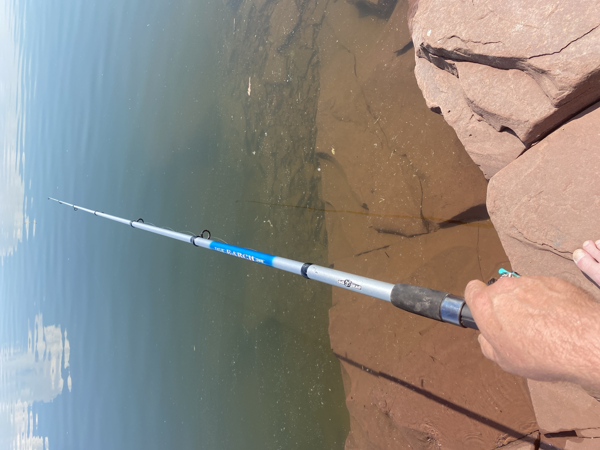 An over-the-shoulder perspective captures a person's hand holding a fishing rod poised over the edge of a body of water, its surface reflecting the sky with wispy clouds. The water, a murky green with reddish-brown sediment visible beneath the surface, laps against a shore composed of large, reddish-brown rocks. The fishing rod, silver with blue accents, extends upwards and to the right, disappearing out of frame. The hand, tanned and wrinkled, grips the black handle of the rod. To the right of the hand, the toes of a bare foot are visible, resting on the rocks. The scene suggests an outdoor activity, likely fishing, in a natural setting characterized by water and rock formations. The location of Ust'-Erba, Russia, is not visually identifiable in the image.