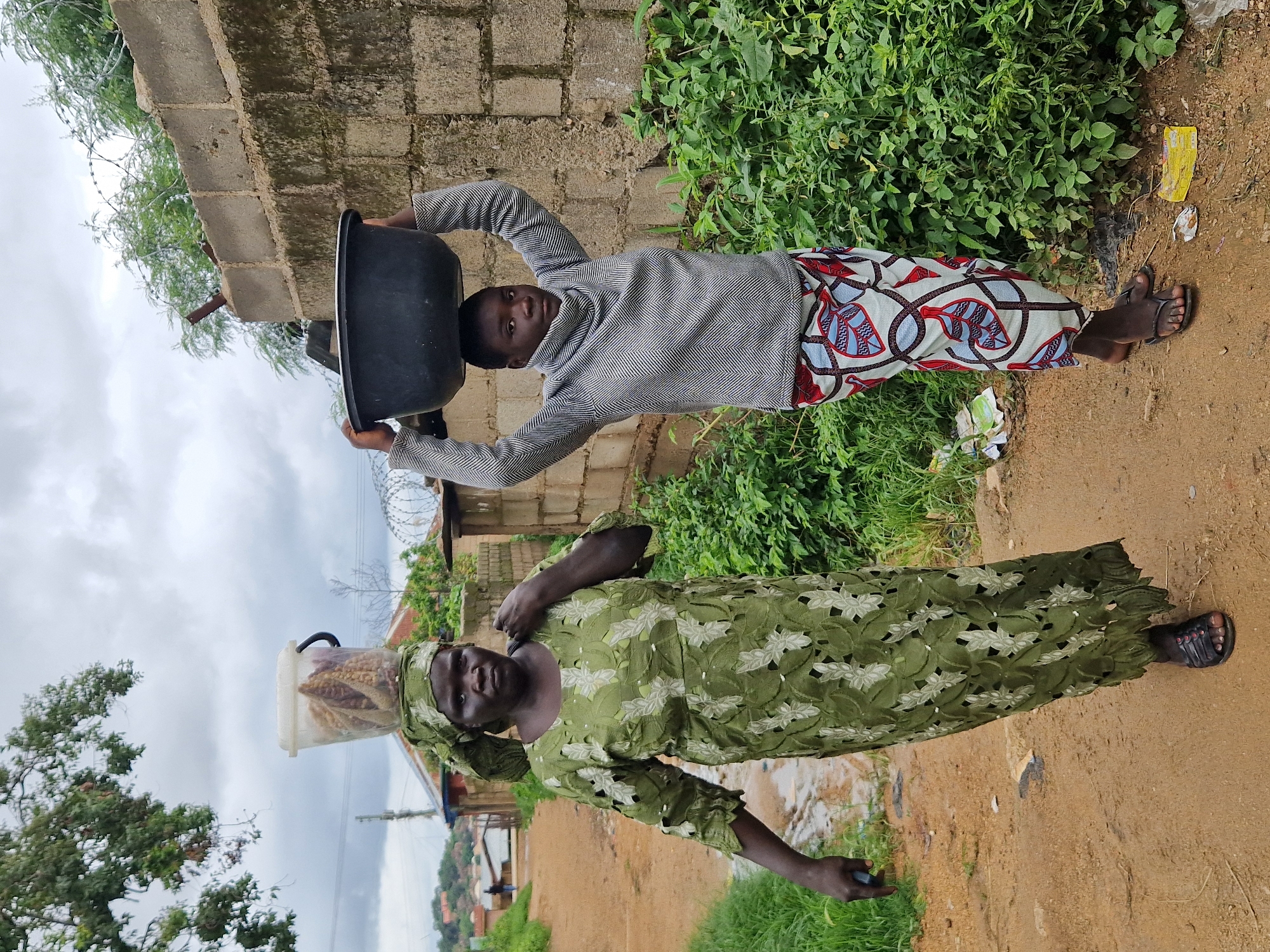 The image captures two individuals on a dirt path in Jos, Nigeria. The person on the left, an adult, walks forward with a bucket containing what appears to be dried corn cobs balanced on their head, covered by a green headscarf. They wear a long, olive-green lace dress with intricate cutouts and black sandals. Their left hand is lowered, holding a dark object. The person on the right, appearing younger, walks forward carrying a large black basin full of water on their head. They are dressed in a grey, high-necked sweater with a chevron pattern and a colorful wrap skirt with a geometric design. They wear dark flip-flops. The environment features a rough, unpaved path with puddles, flanked by dense green foliage on the right. A partially constructed brick wall with barbed wire fencing runs along the right side of the path. In the background, more buildings and greenery are visible, under an overcast sky.